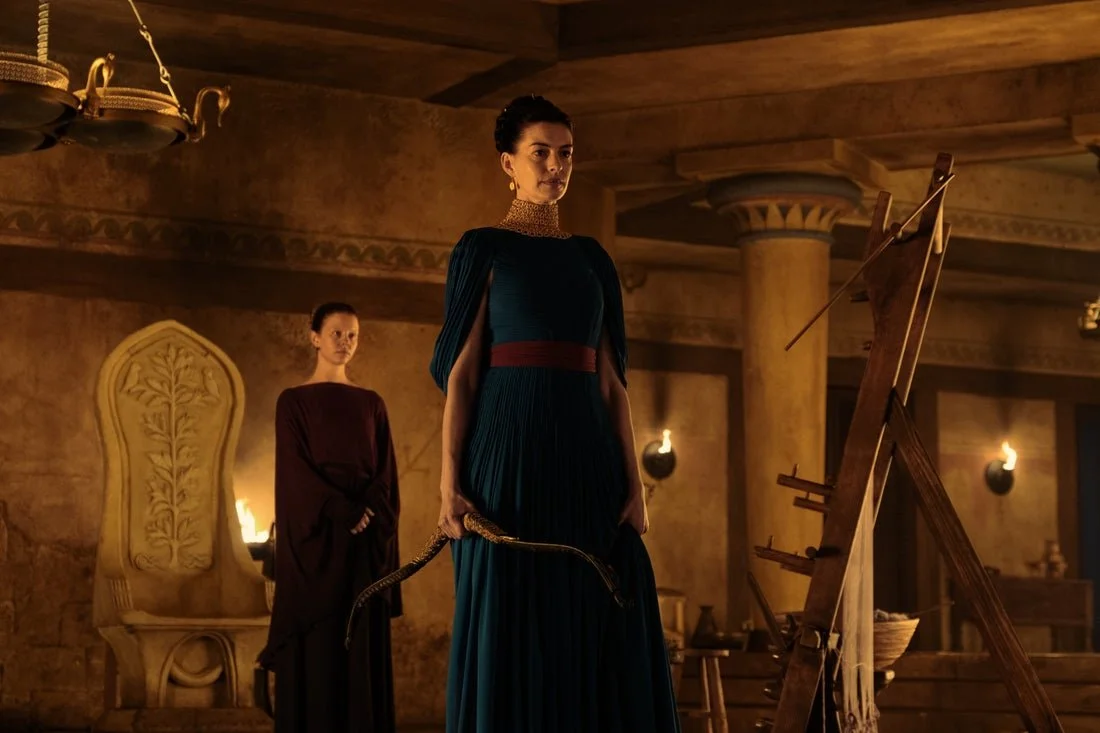In Christopher Nolan's film adaptation of THE ODYSSEY, Wise Queen Penelope (Anne Hathaway) of Ithaca holds Odysseus' famed bow in front of her loom, her servant Melantho (Mia Goth), and her own throne.