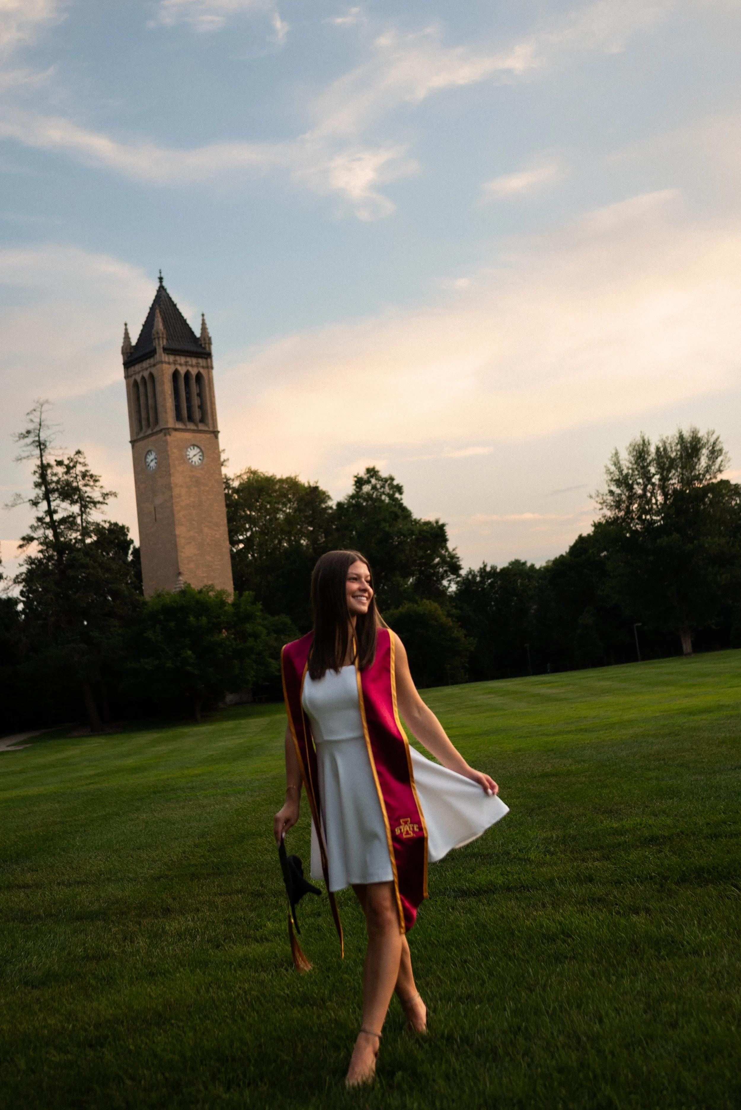 A young woman in a white dress and a graduation stole is walking on a grassy area near a tall clock tower and trees, smiling, at sunset or late afternoon.