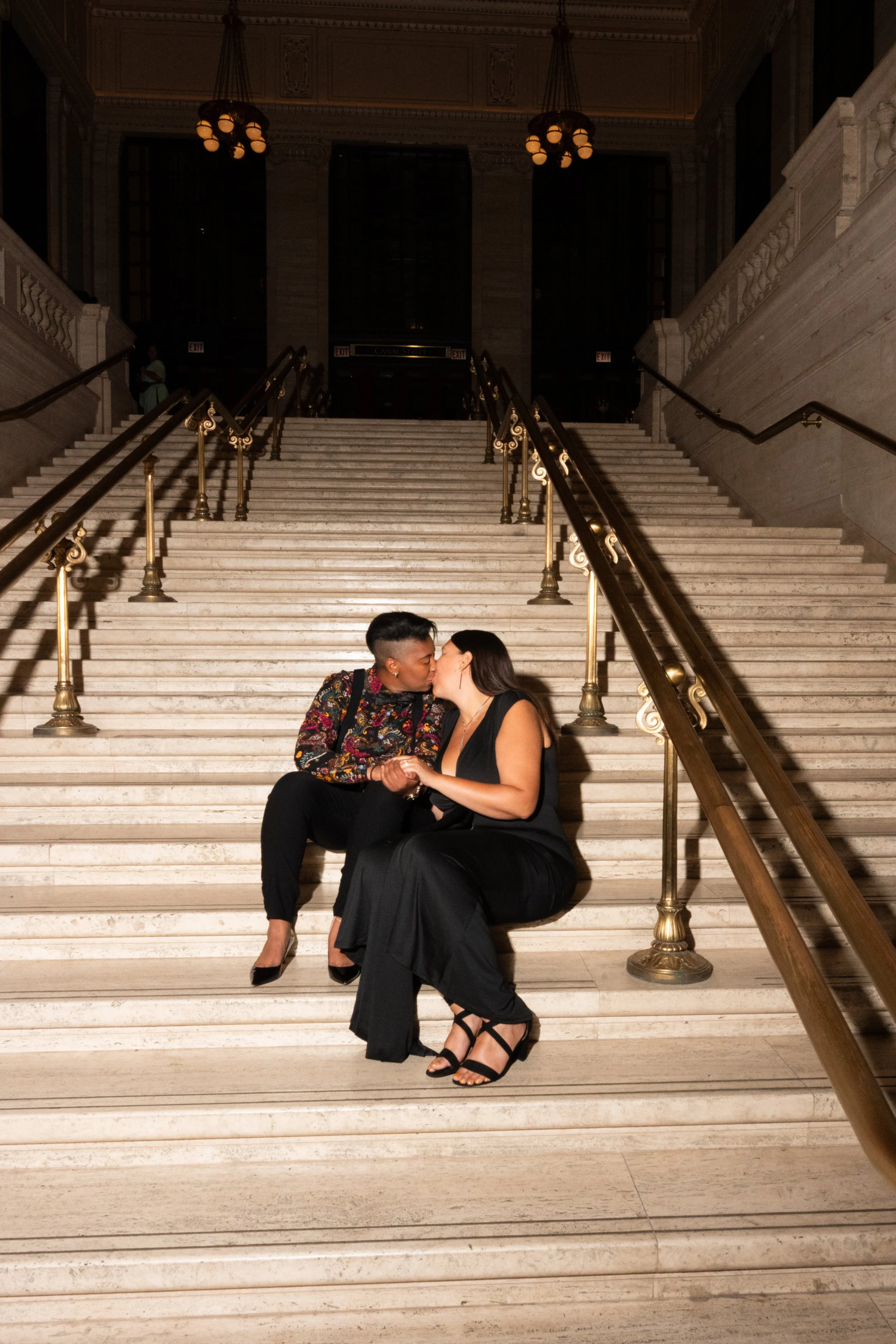 A couple is sitting on the stairs inside a grand building, sharing a kiss, with the woman wearing a black gown and the man wearing a colorful patterned jacket, both dressed elegantly.