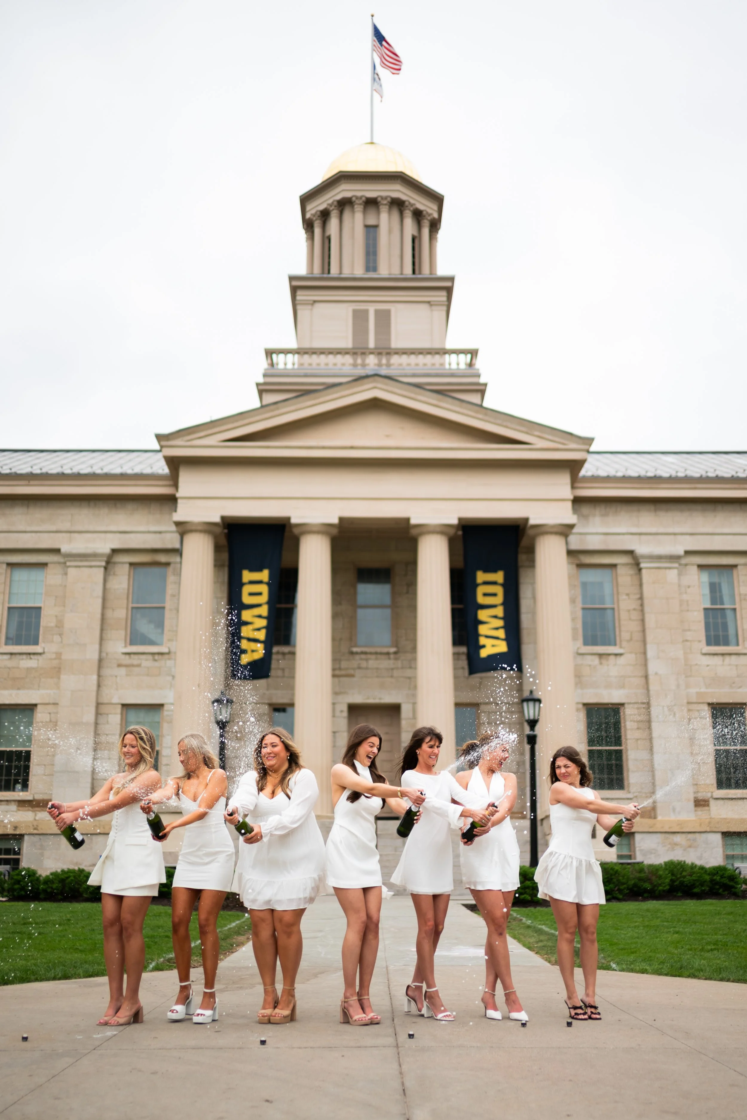 Seven women in white dresses celebrating with Champagne bottles on a sidewalk in front of a courthouse, with fountains spraying water on each woman, and an American flag on top of the courthouse.
