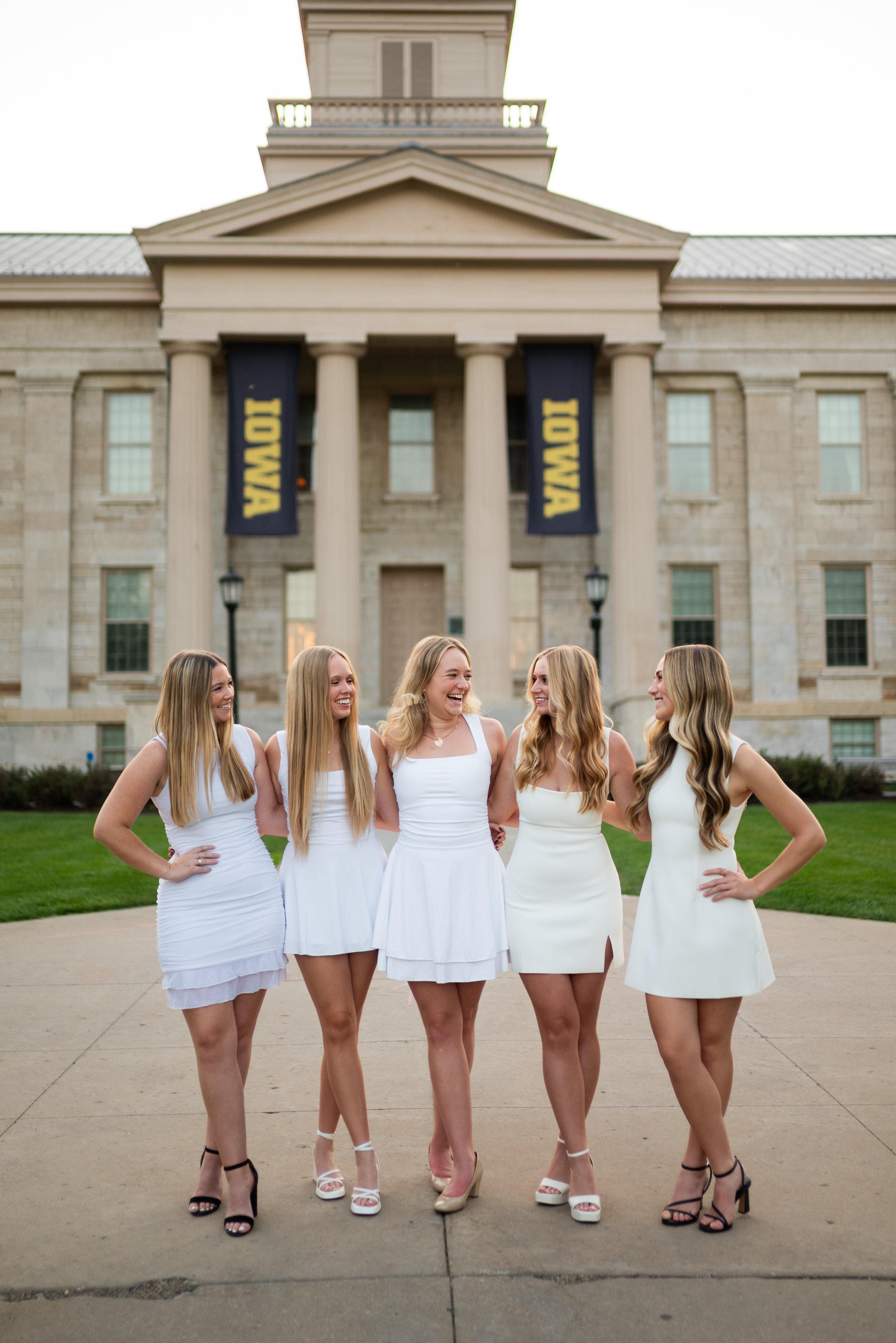 Five women dressed in white dresses standing together in front of a large building with columns and banners that read 'IOWA', smiling and enjoying each other's company.