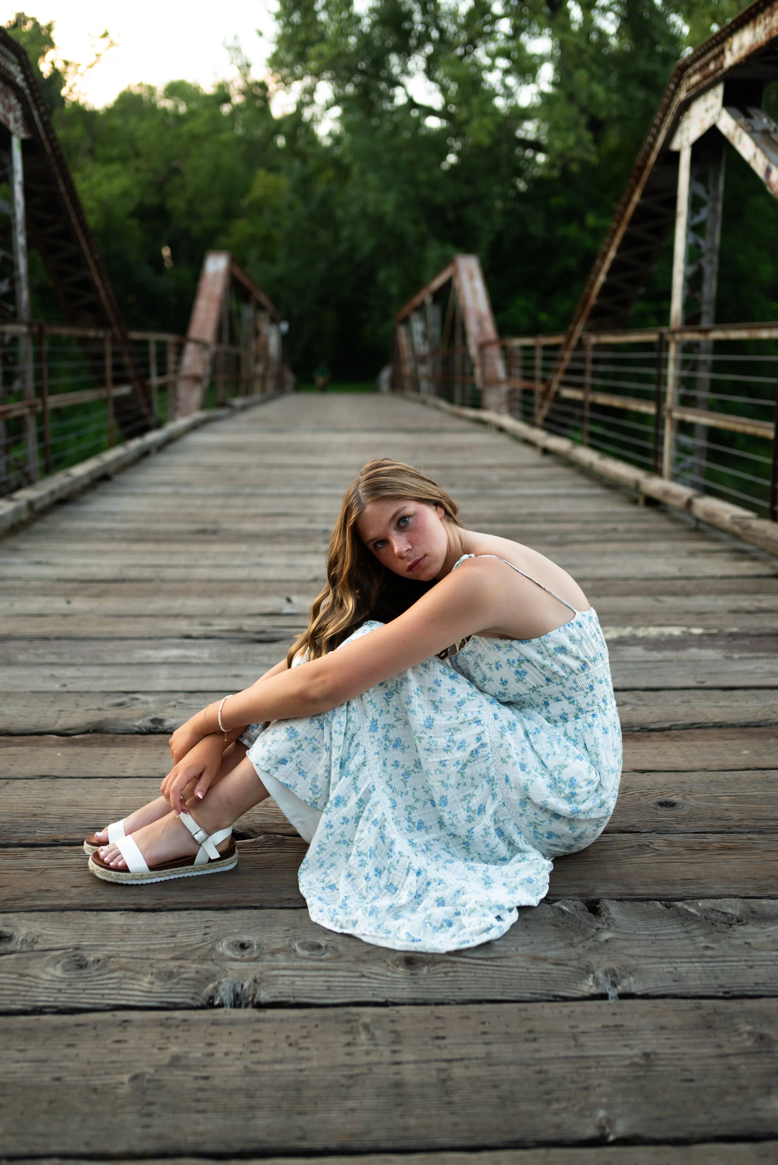 Young woman sitting on a wooden bridge wearing a blue floral dress and white sandals with a contemplative expression.