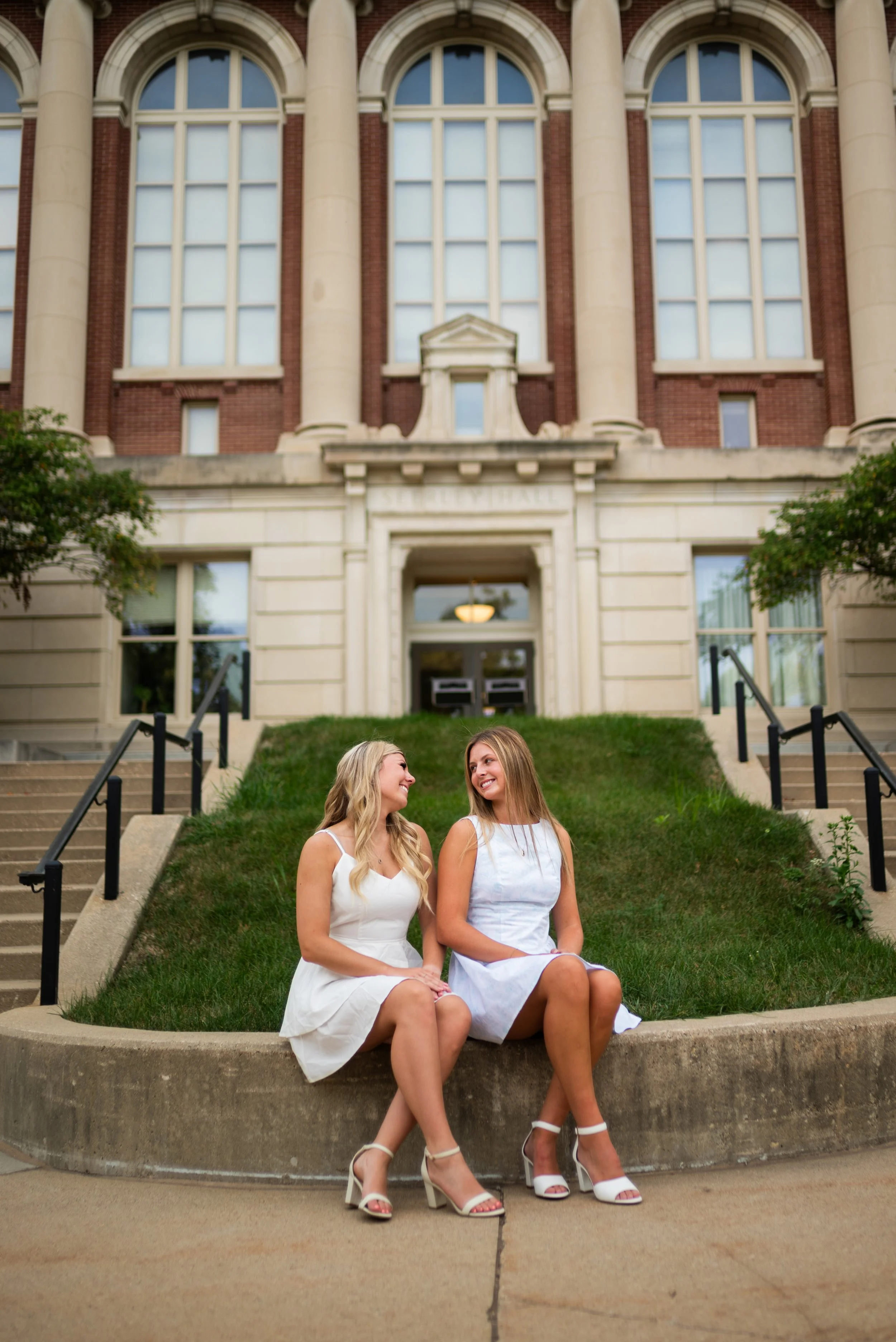 Two young women in white dresses and high heels sitting on a concrete ledge in front of a historic brick building with large arched windows, smiling at each other.