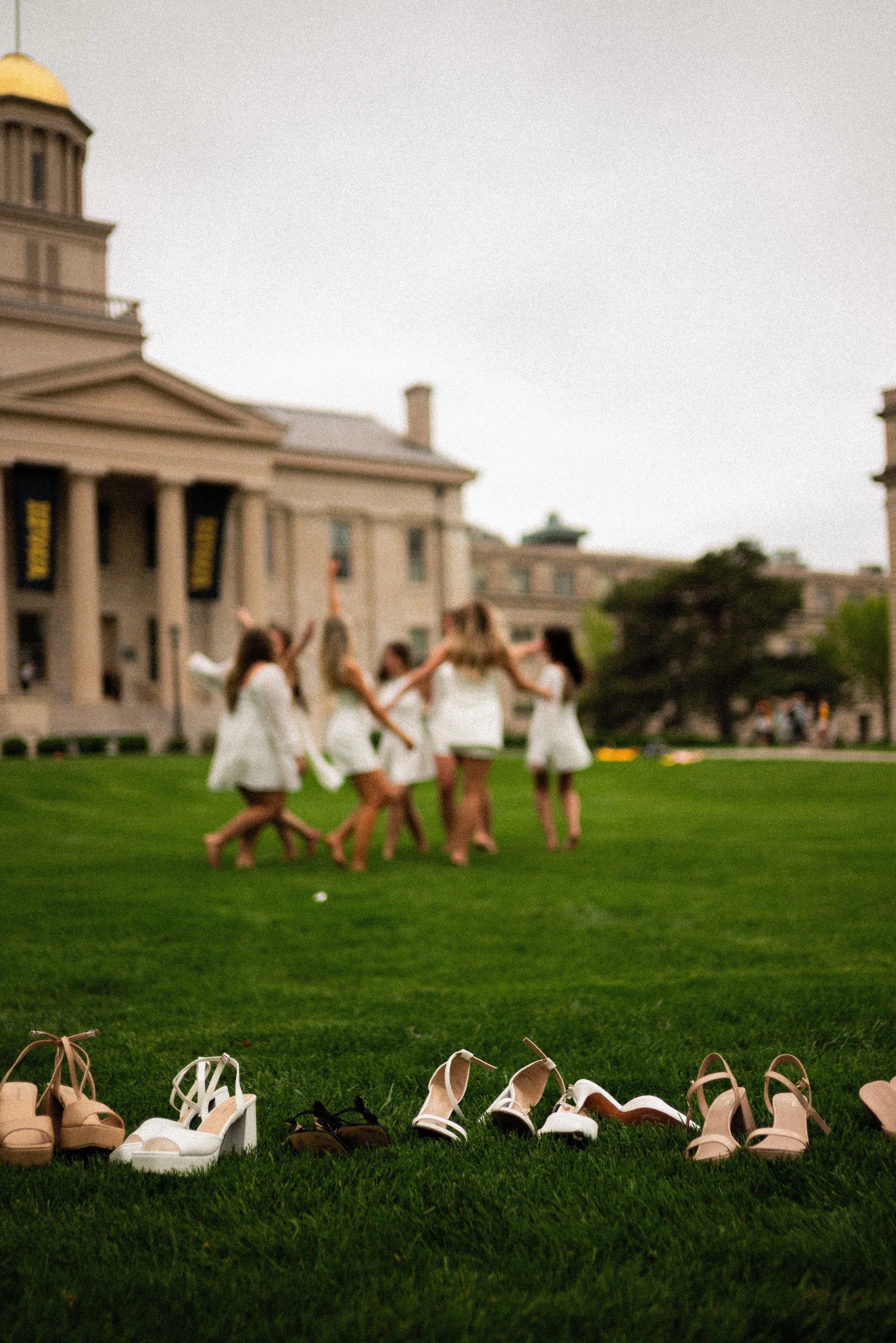 A row of various high-heeled shoes on green grass, with a group of women in white dresses dancing in the background near a large historic building with columns.