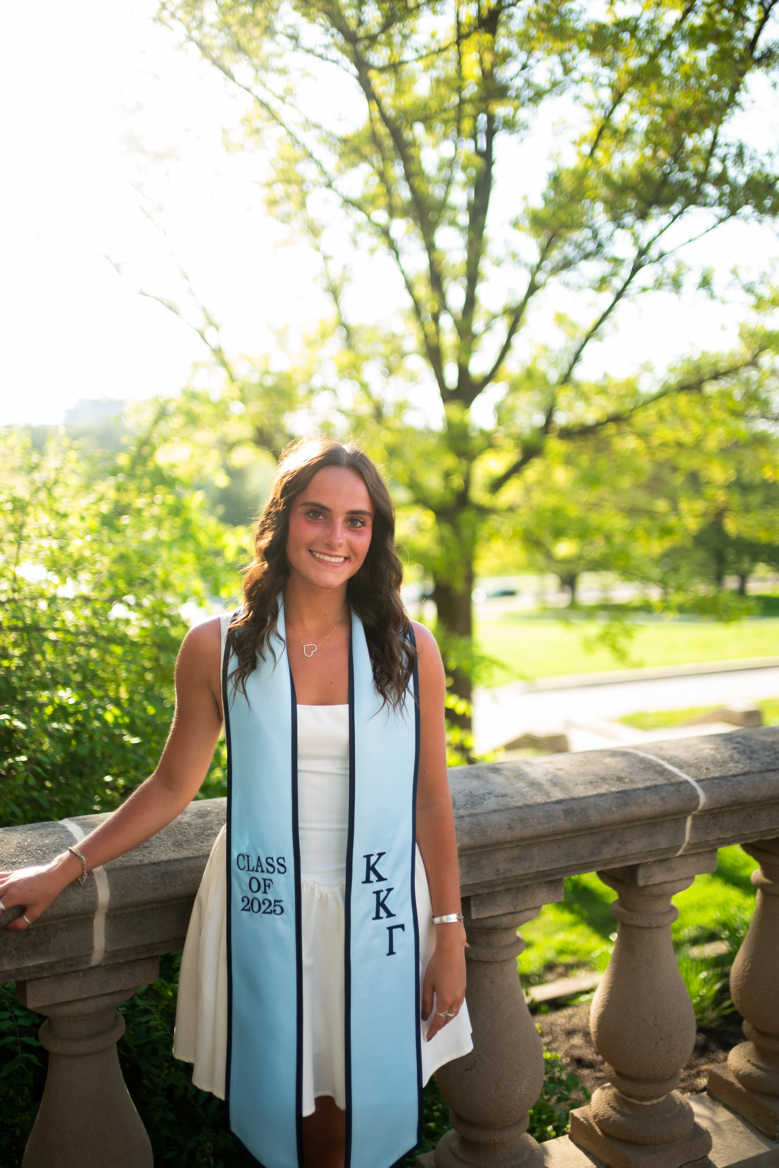 A young woman standing outdoors on a stone railing, wearing a white dress and a light blue graduation stole with the words "Class of 2025" and Greek letters, smiling at the camera with trees and greenery in the background.