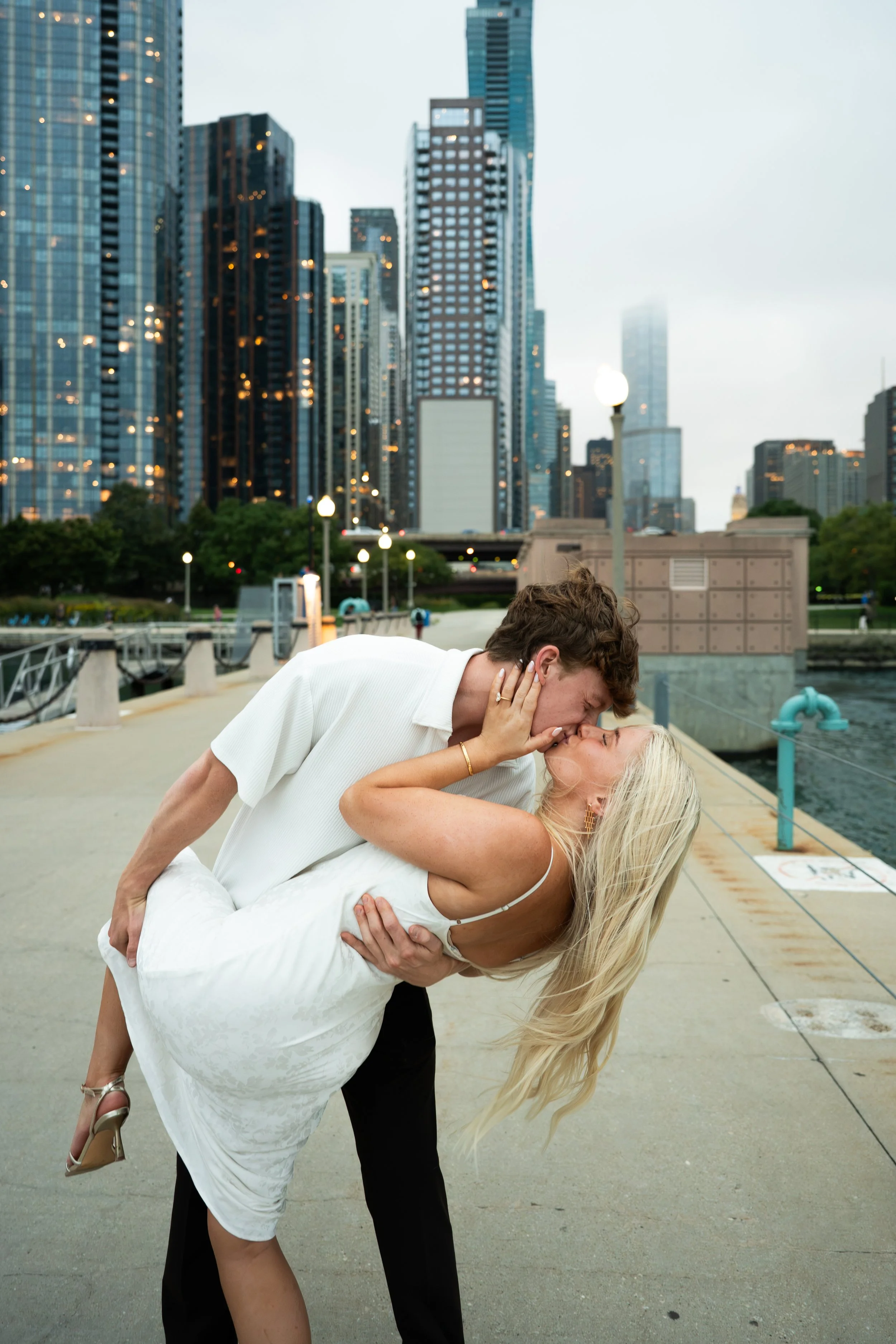 A man and woman kiss on a city sidewalk, with the man leaning over and holding the woman, who is wearing a white dress. The woman has blonde hair and is wearing high heels. The background features tall modern skyscrapers and city lights.