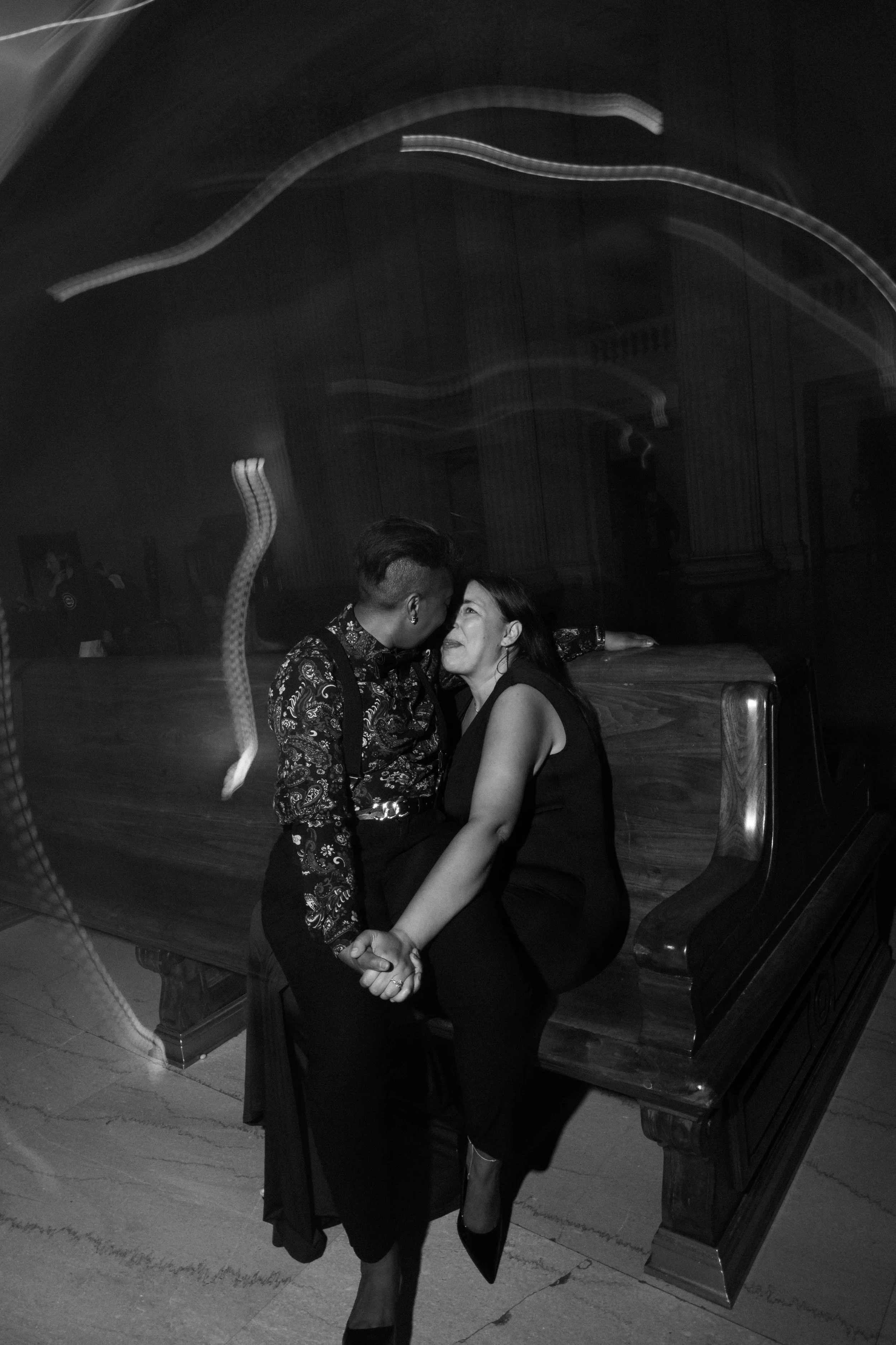 A black and white photo of a couple dancing closely together in a dimly lit room, with a vintage wooden bench and ornate interior details in the background.