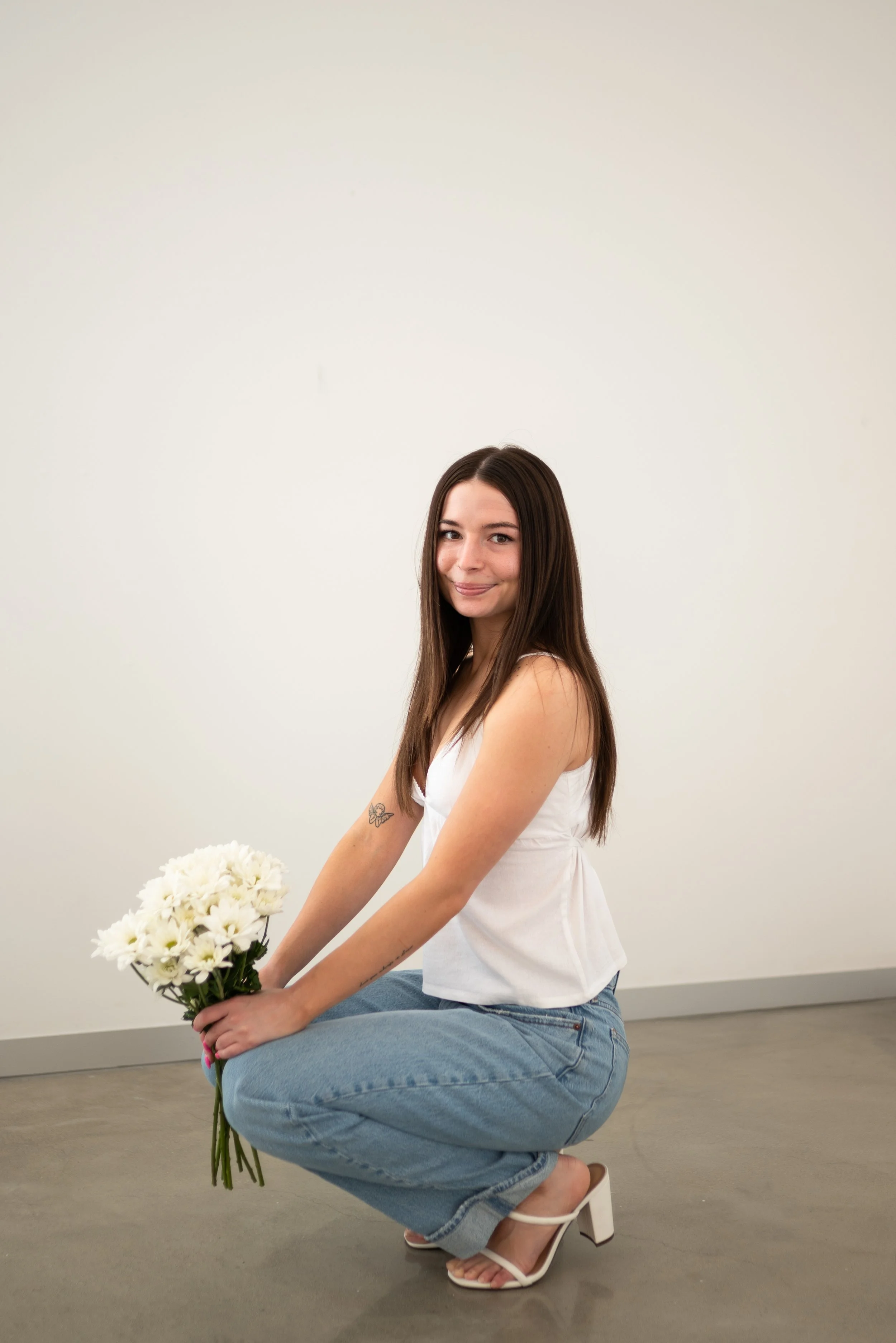A woman with long brown hair, smiling, holding a bouquet of white flowers, crouching on the floor against a plain white wall, wearing a white sleeveless top, light blue jeans, and white high-heeled sandals.