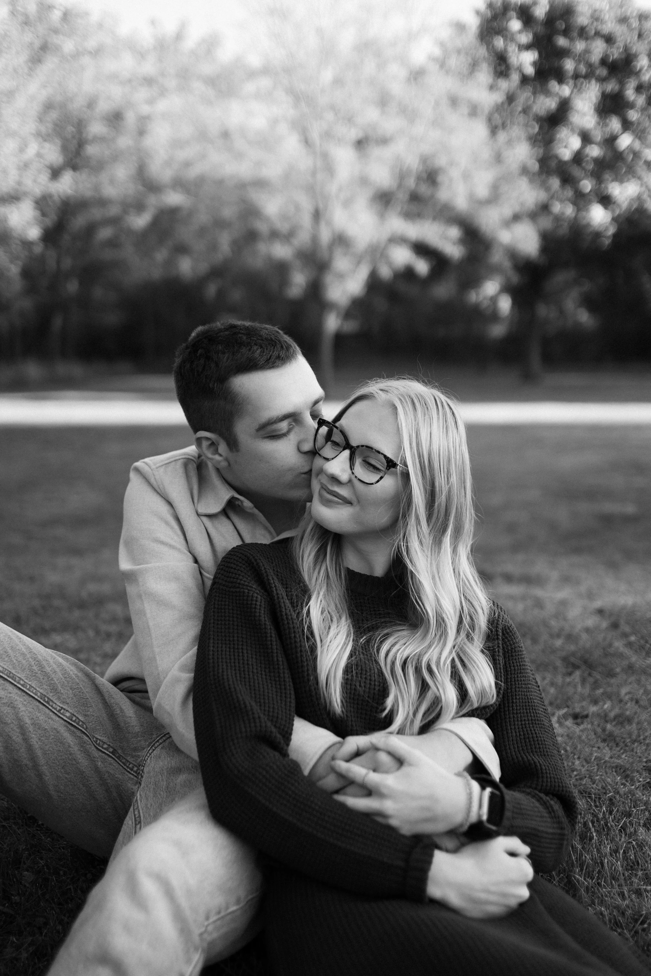Young couple sitting on grass in a park, the man kissing the woman on the cheek, the woman smiling with eyes closed, in black and white.