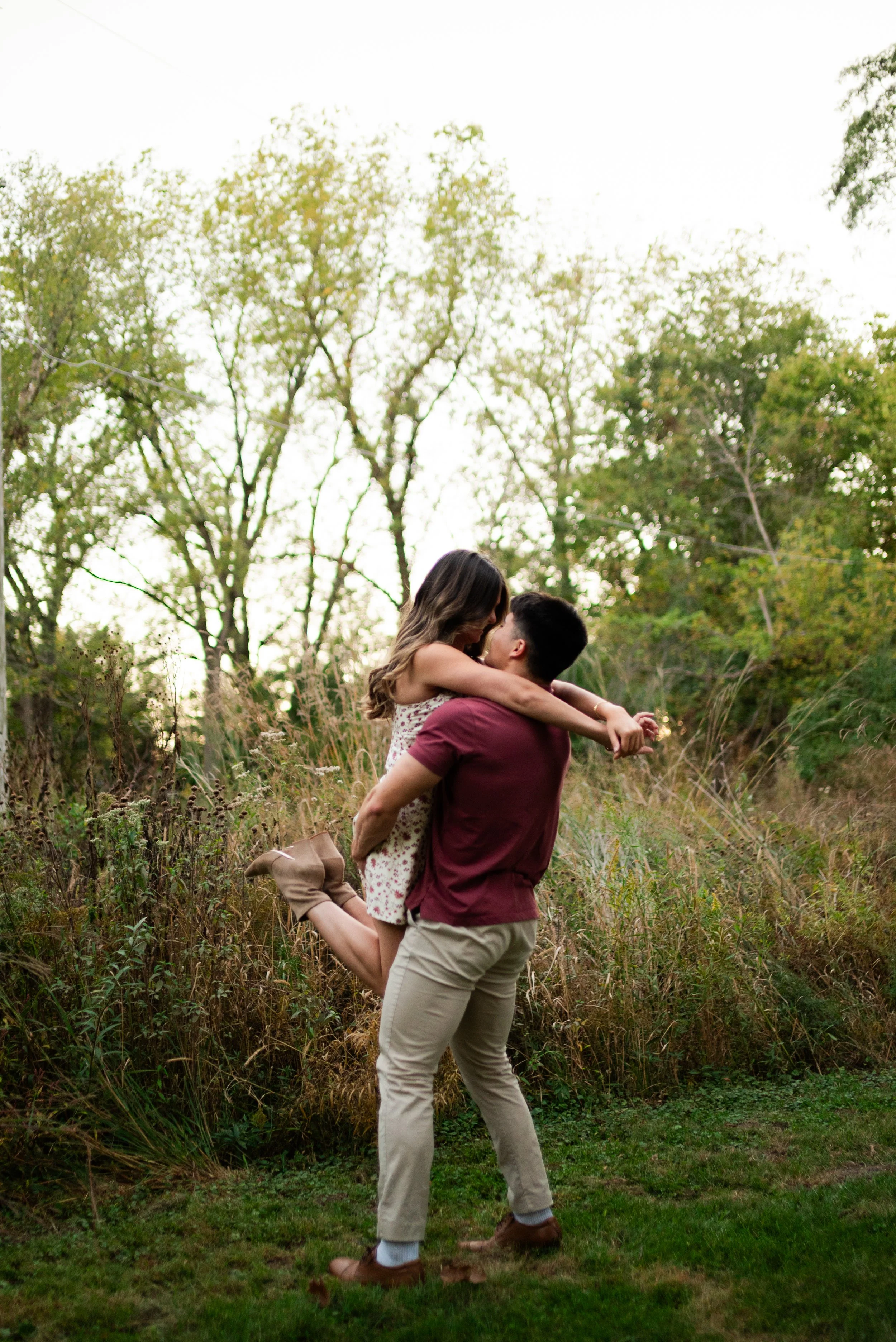A man lifts a woman in his arms outdoors during daytime, surrounded by trees and grass, as they share a romantic moment.