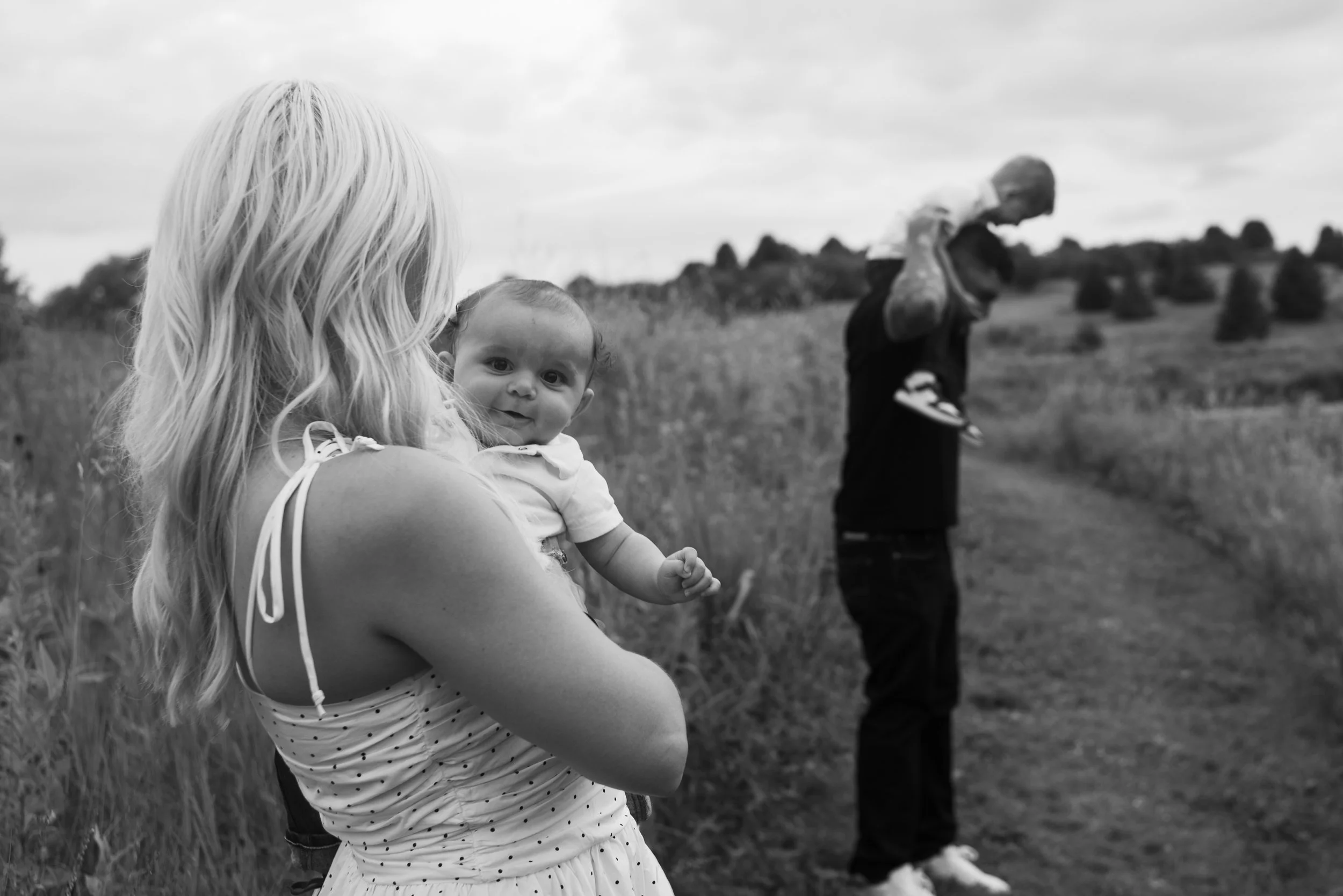 A woman holding a baby in a field, with a man in the background carrying a child on his shoulders.