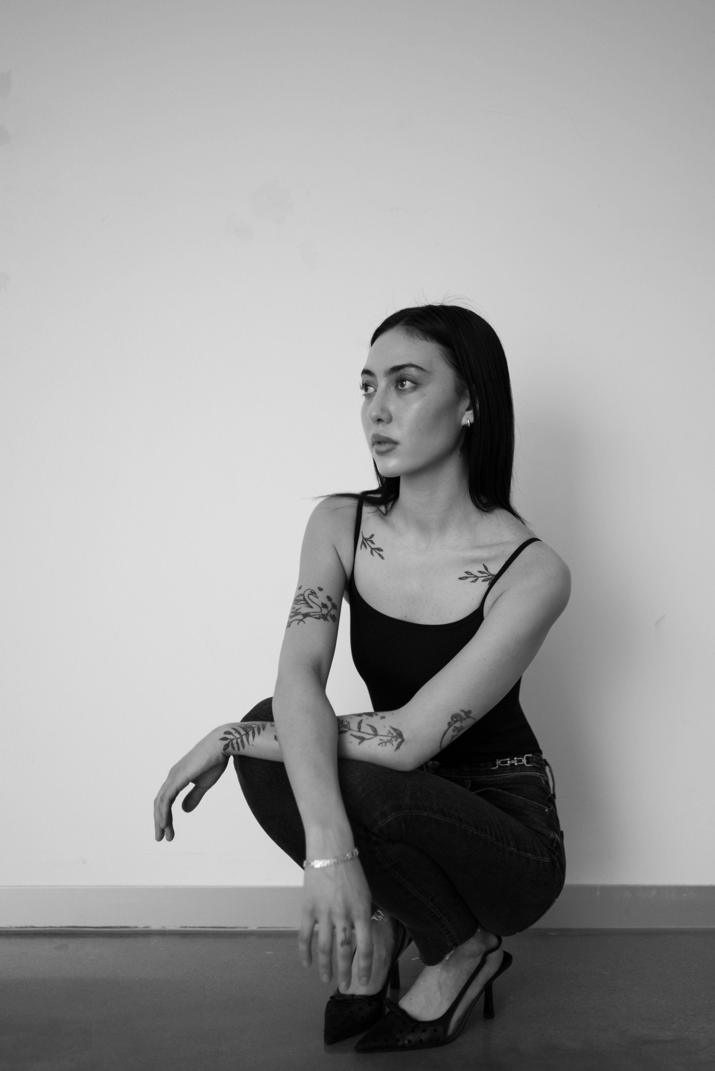 A black and white photo of a woman with dark hair, tattoos on her arms and shoulders, wearing a black tank top, black jeans, and high heels, crouching against a plain white wall, looking to her left.