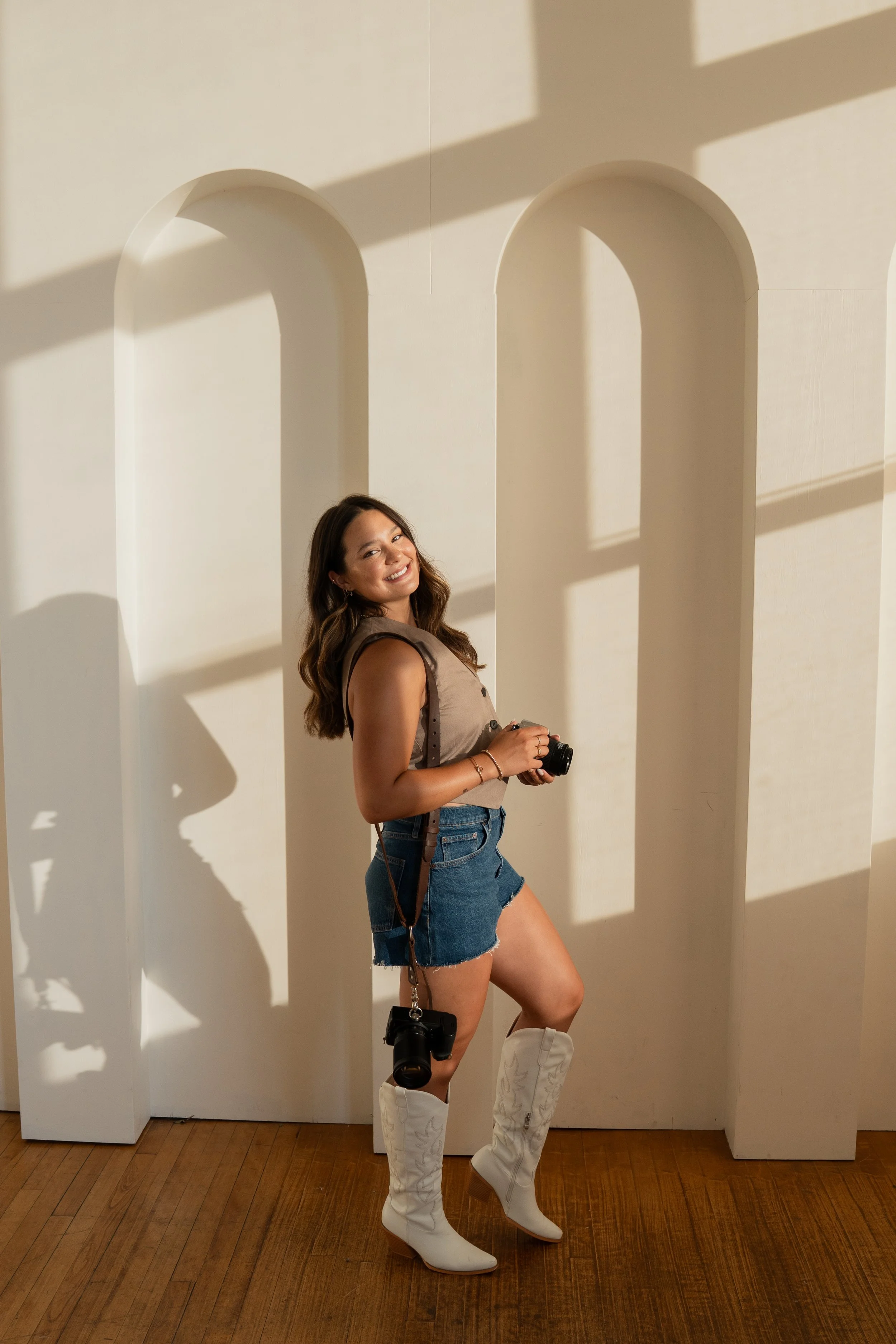 A young woman standing indoors on a wooden floor, smiling, holding a camera, wearing a sleeveless top, denim shorts, and white cowboy boots, with shadows cast on the wall behind her.