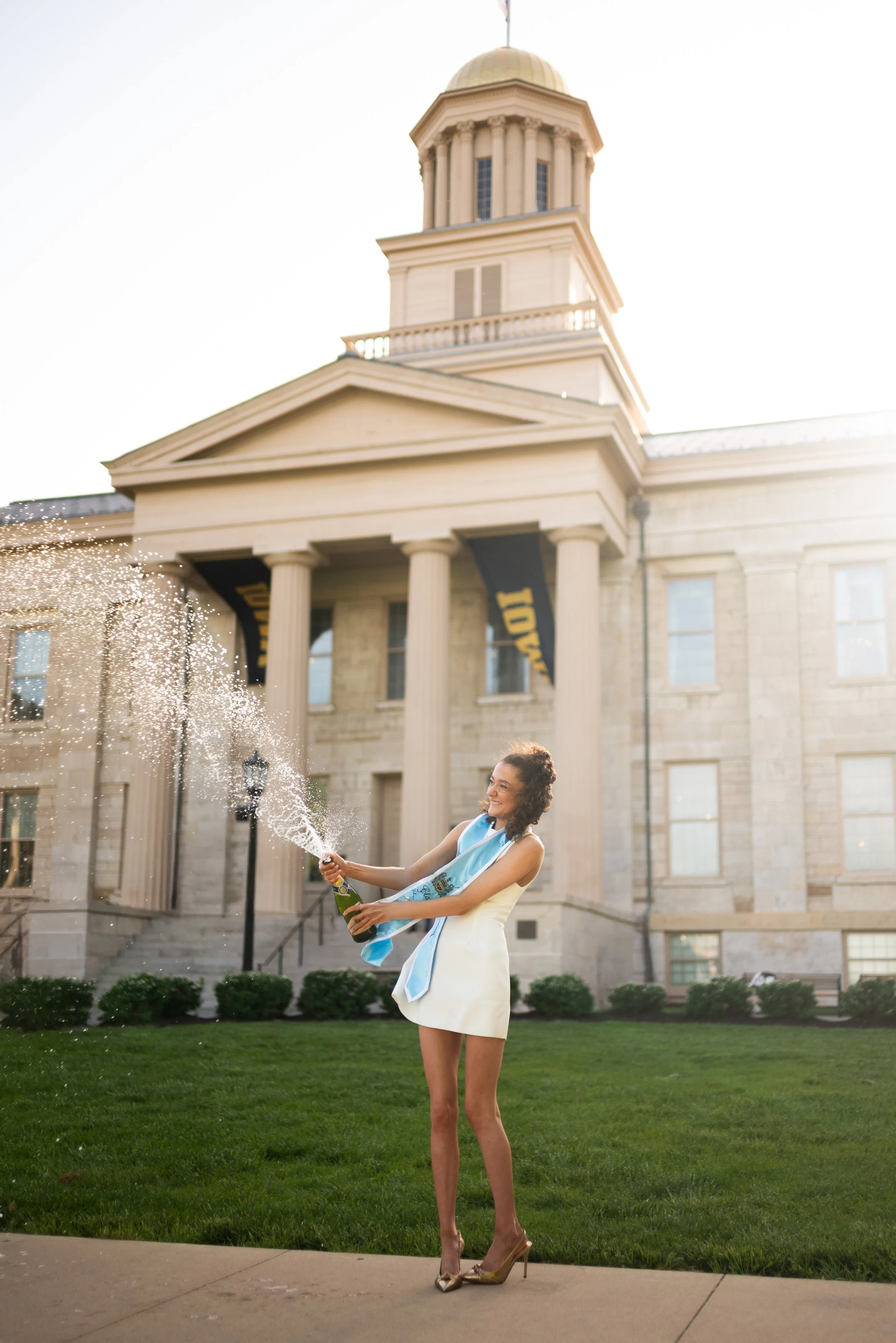 A woman in a white dress and high heels celebrating graduation with a champagne bottle and burst of champagne in front of a historic courthouse building.