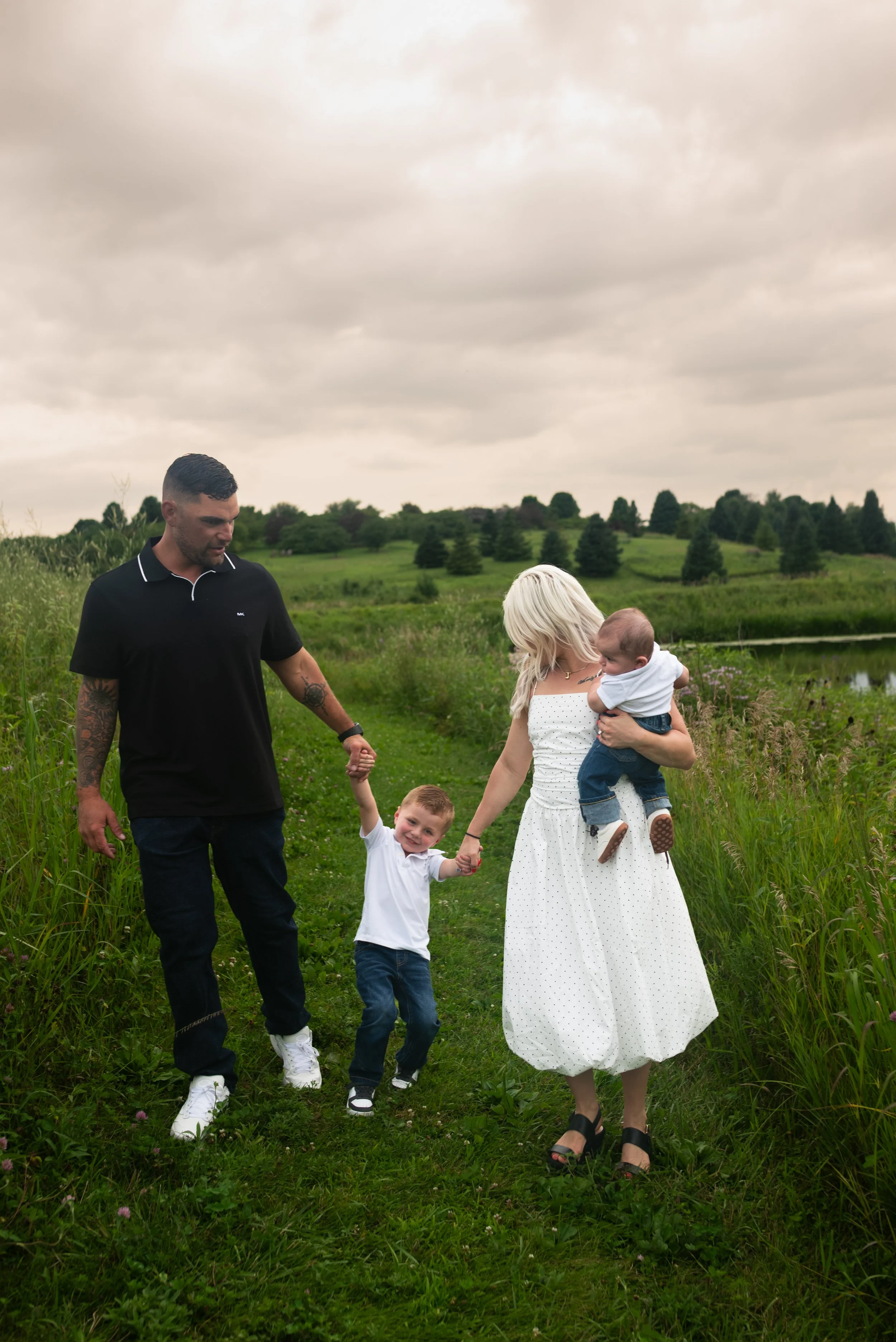 Family walking on a grassy path outdoors, with a man, woman, and two children, in a rural setting with green fields and trees under a cloudy sky.