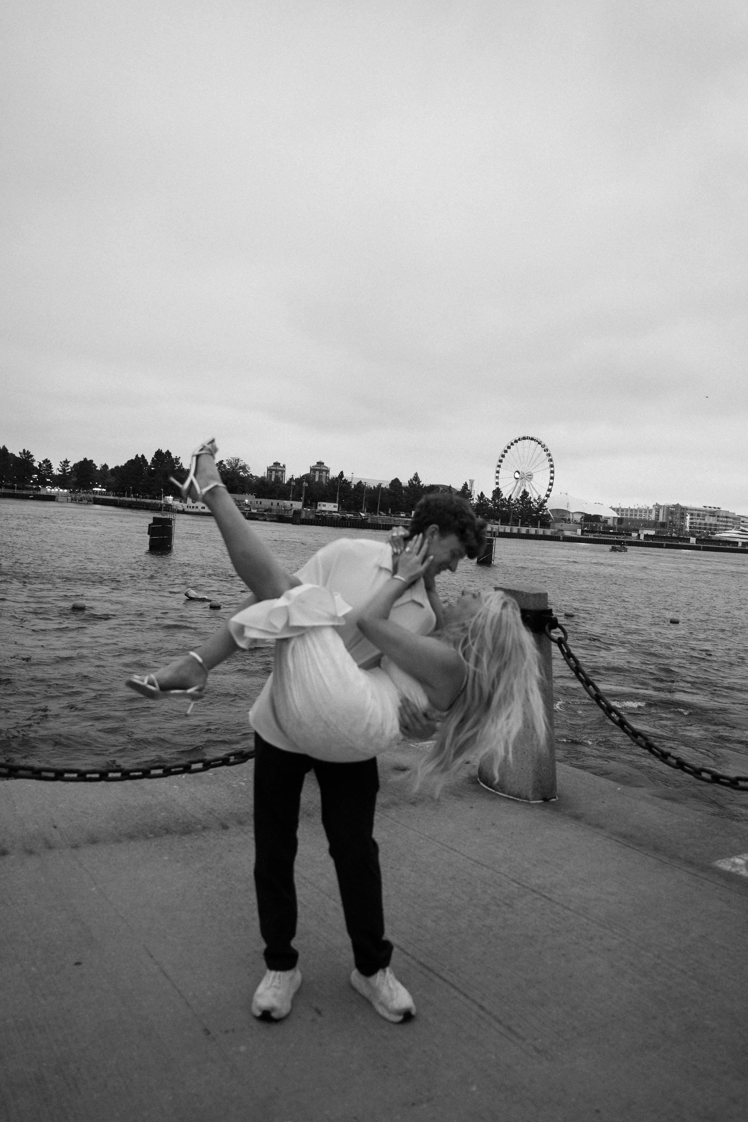 A man is holding a woman upside down by her waist and her head near the ground near a waterfront, with a Ferris wheel and city buildings in the background.