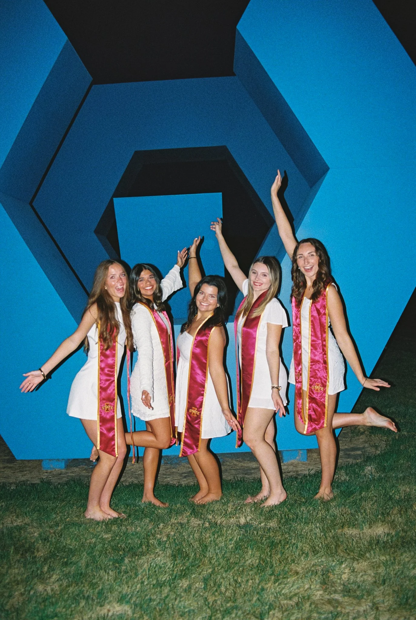 Group of six young women wearing graduation stoles and dresses, celebrating outdoors at night in front of a large, blue, geometric art installation.