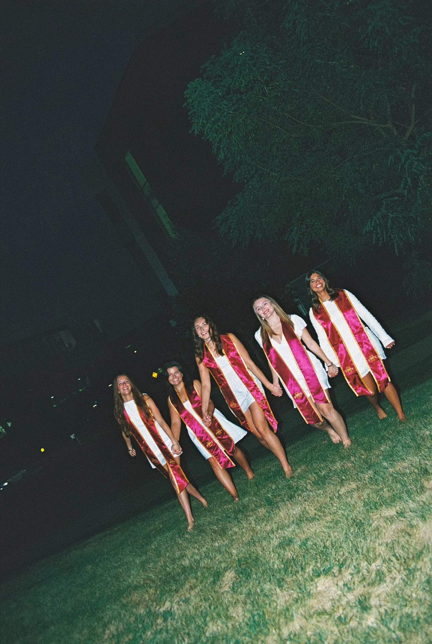 Five young women in graduation gowns holding hands and smiling outside at night.