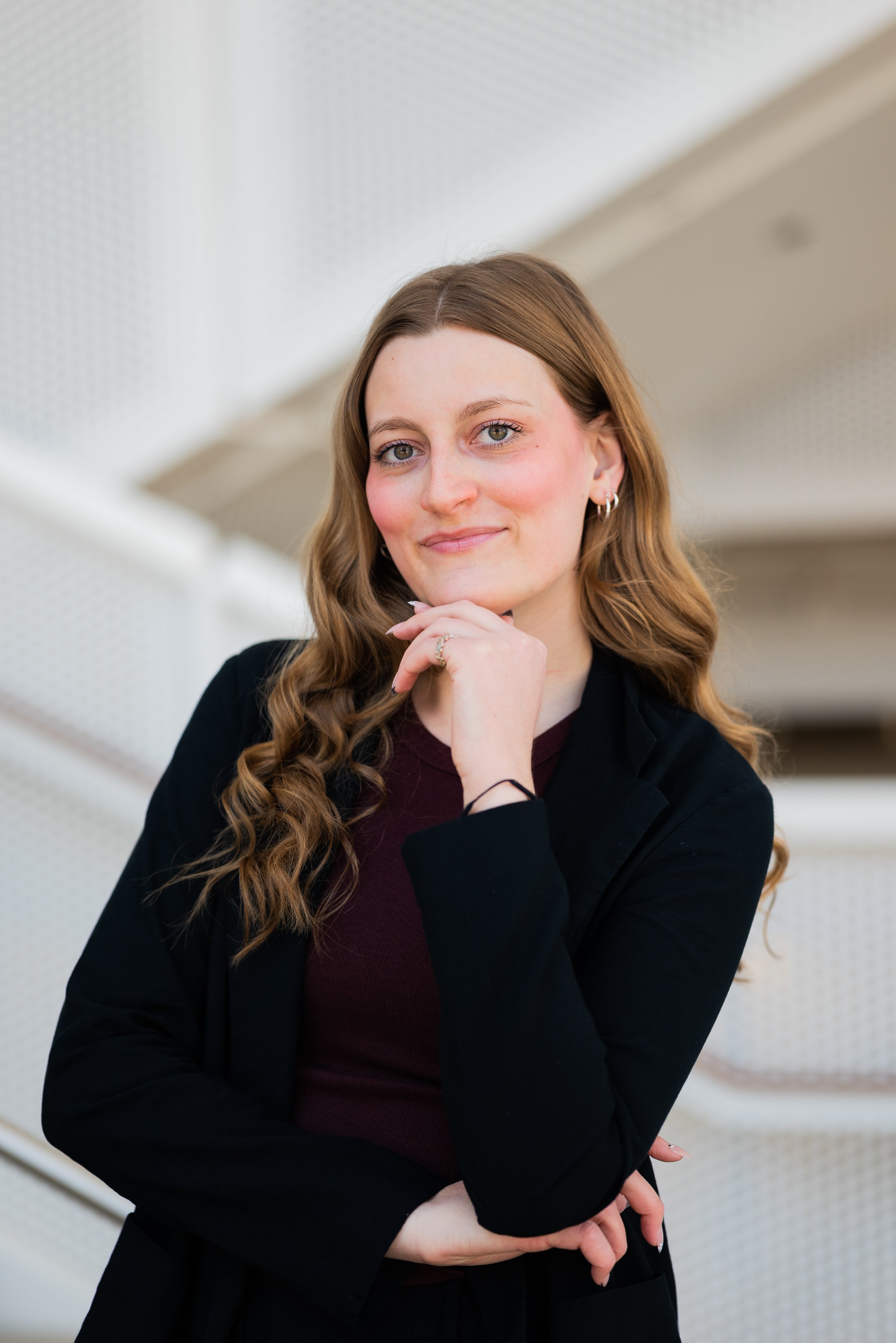 A young woman with long wavy brown hair, wearing a black blazer and maroon shirt, stands indoors with her hand on her chin, smiling softly at the camera.