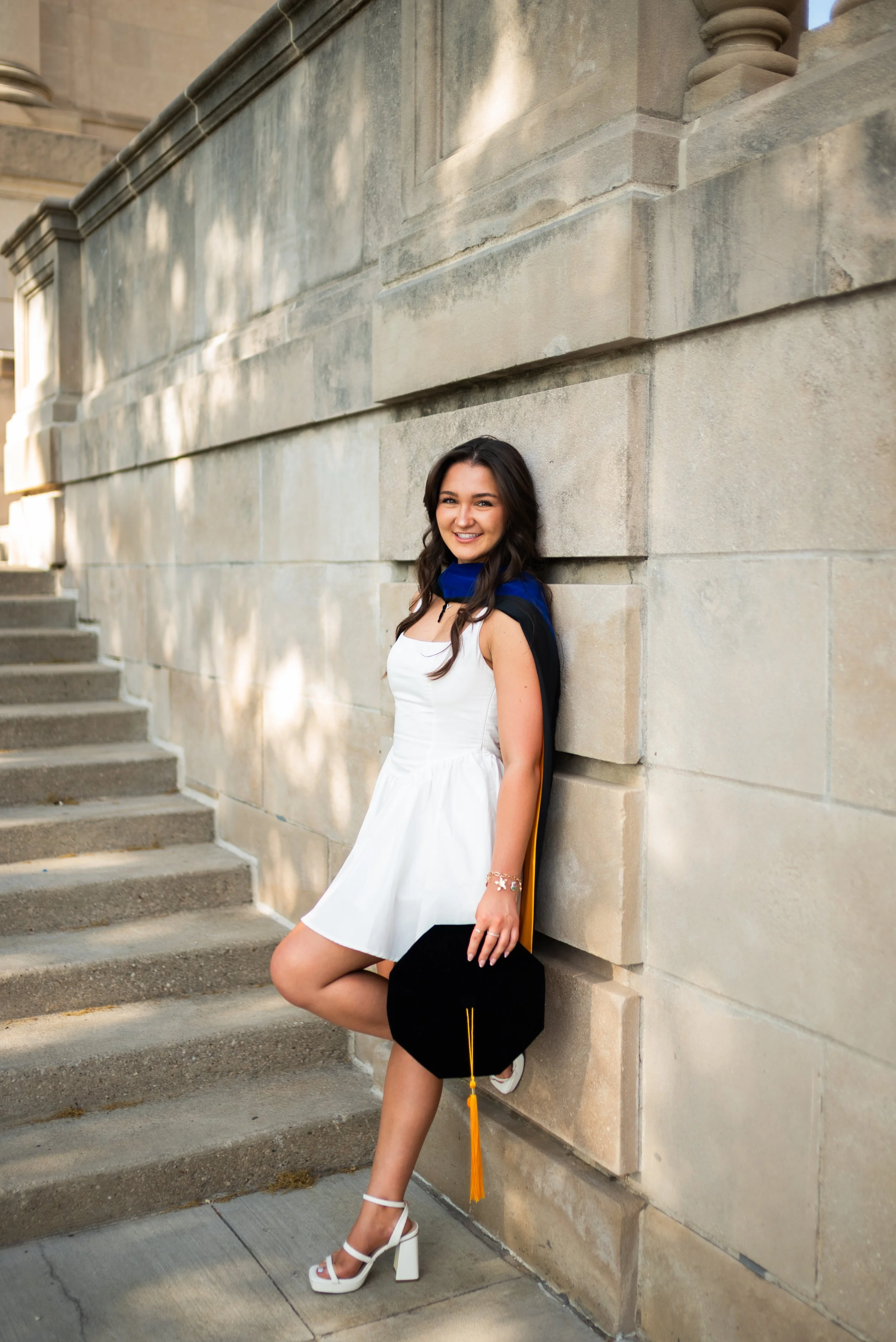 Young woman in a white dress with a blue and black graduation stole, standing against a stone wall holding a black graduation cap with a yellow tassel, smiling, outside on steps.