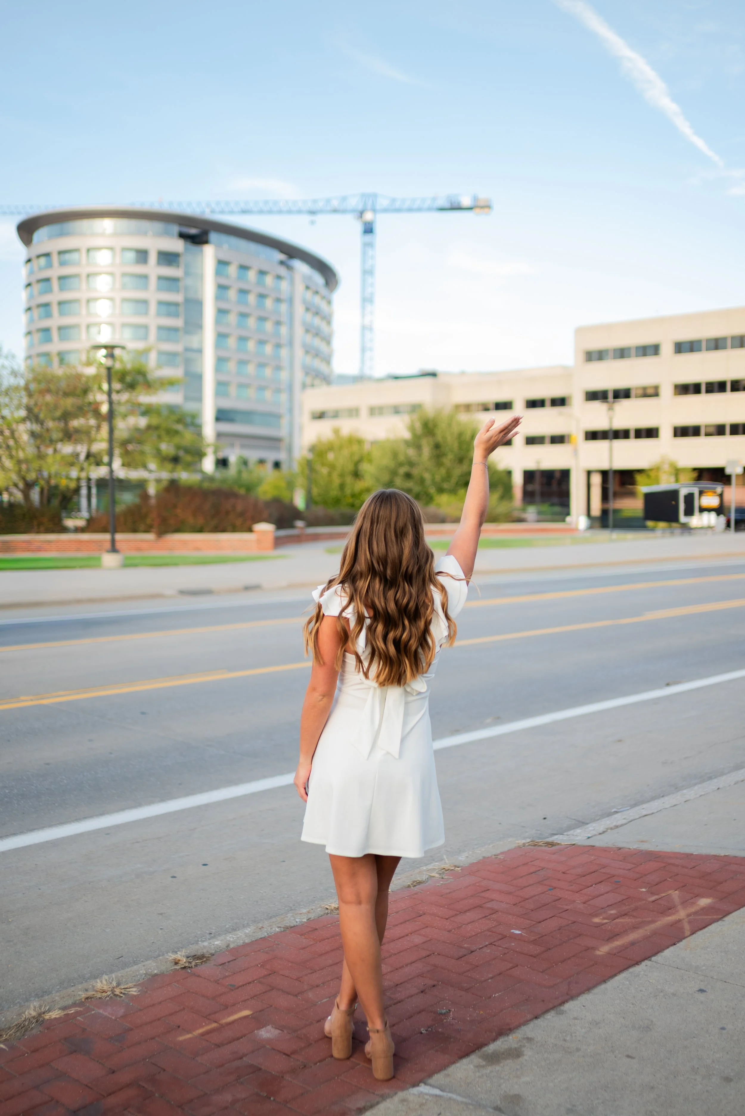 A woman with long, wavy brown hair wearing a white dress and beige high heels standing on a sidewalk, waving to a building across the street on a sunny day.