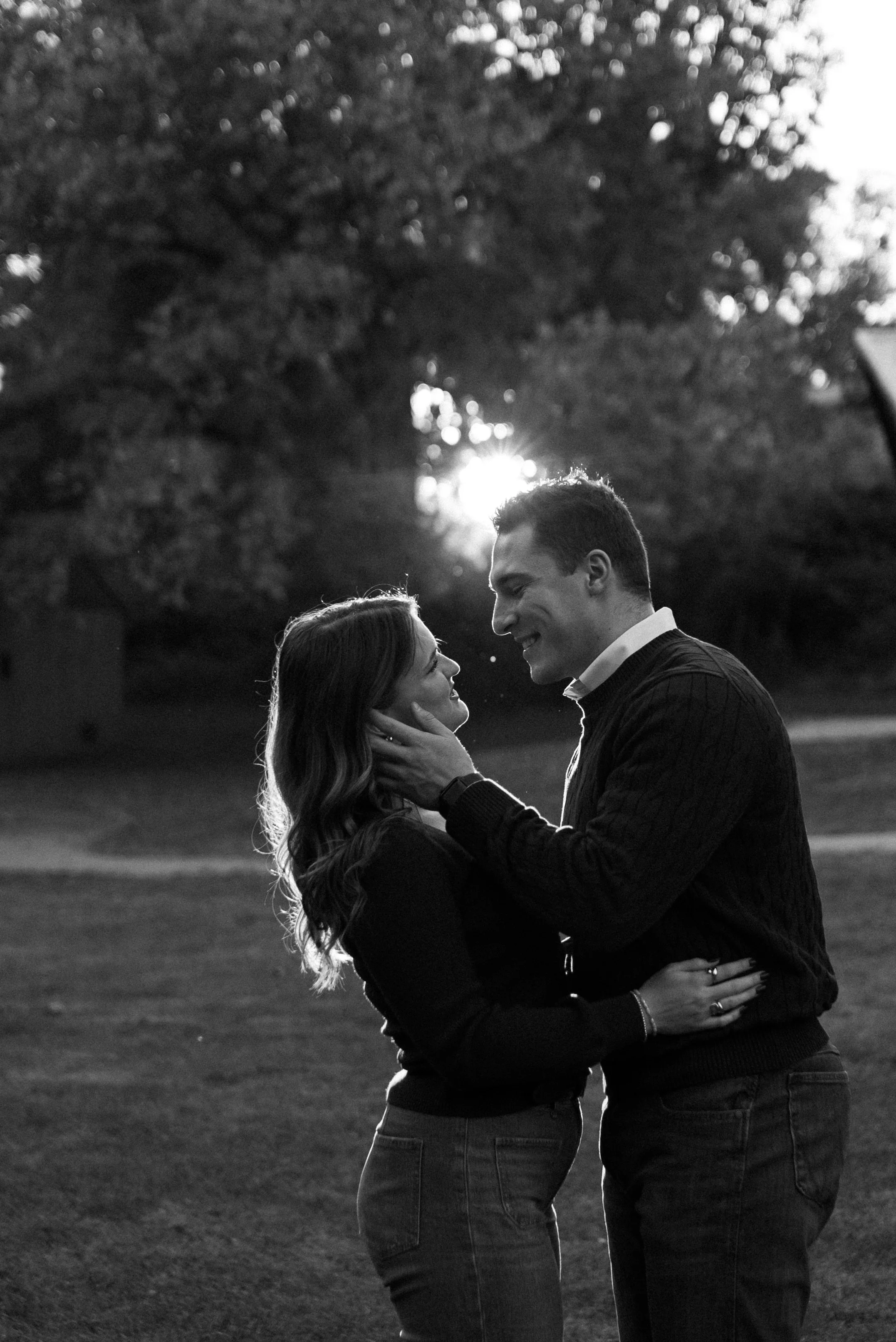 A black and white photo of a couple standing close together outdoors during sunset. The man gently holds the woman's face with one hand, and they are gazing into each other's eyes with smiles. Trees and the sun are visible in the background.