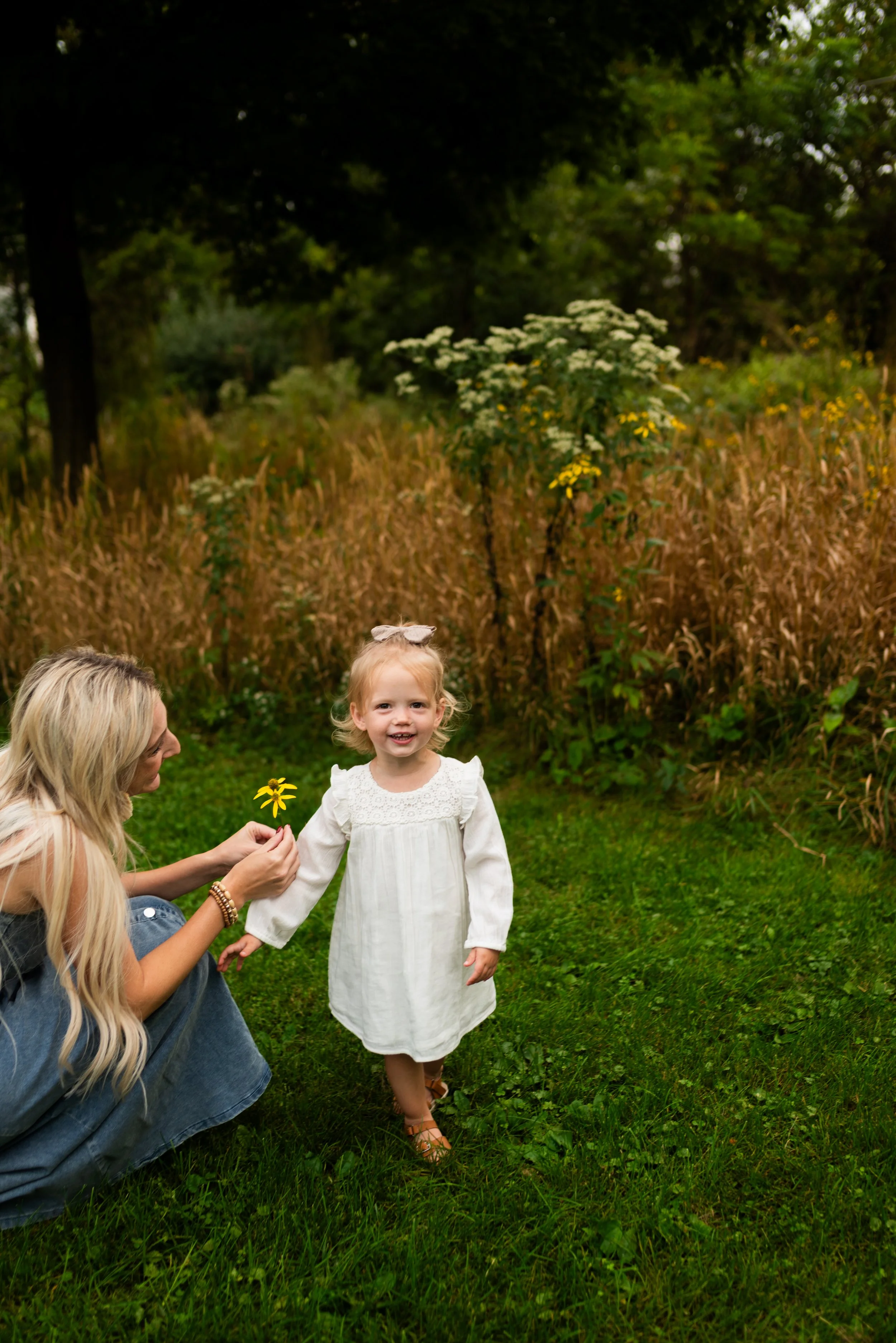 A woman kneeling on the grass holding a yellow flower, smiling at a young girl in a white dress, outdoors in a natural setting with trees and tall grass in the background.