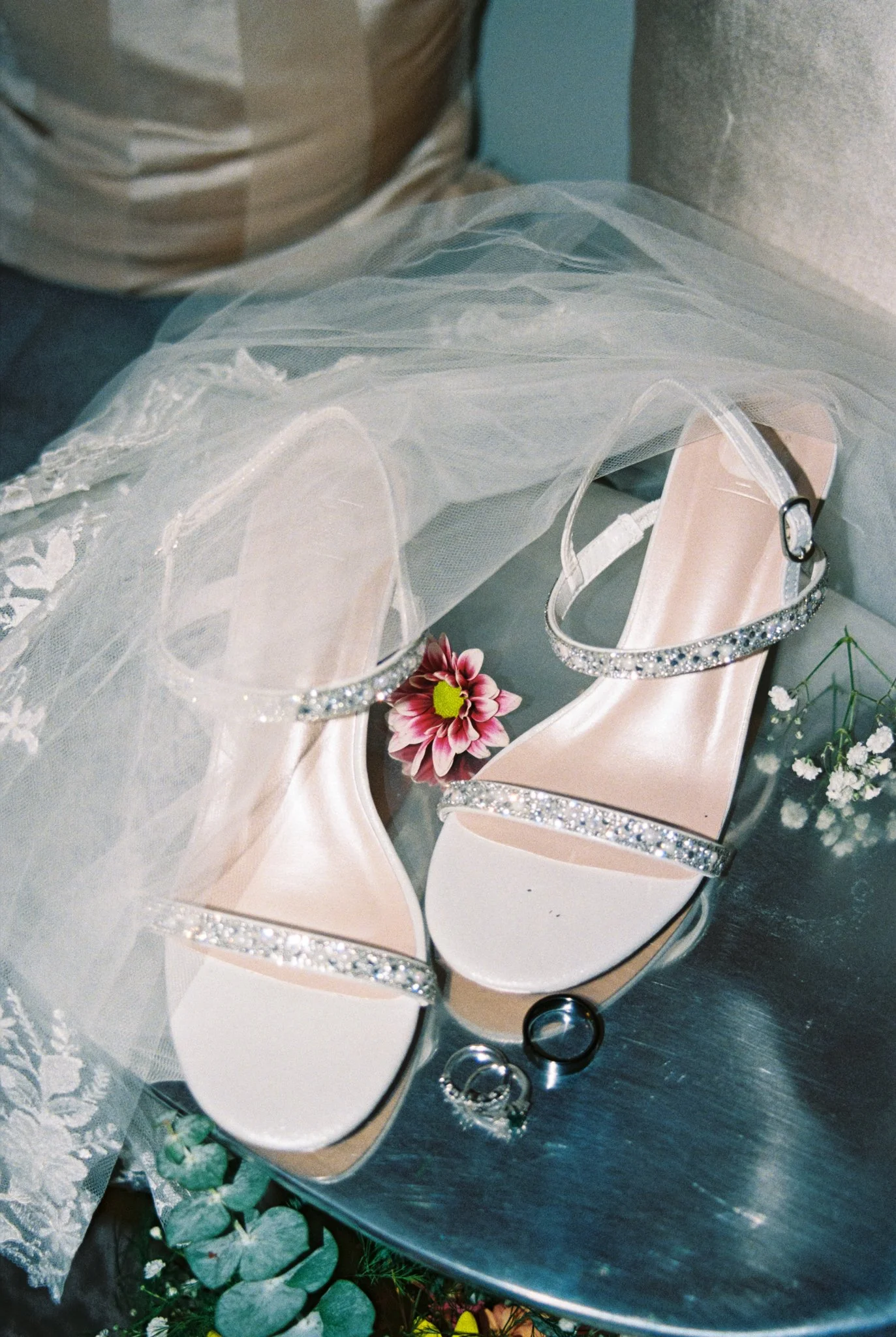A pair of white wedding shoes with rhinestone straps, a veil, a pink flower, and wedding rings on a silver surface.