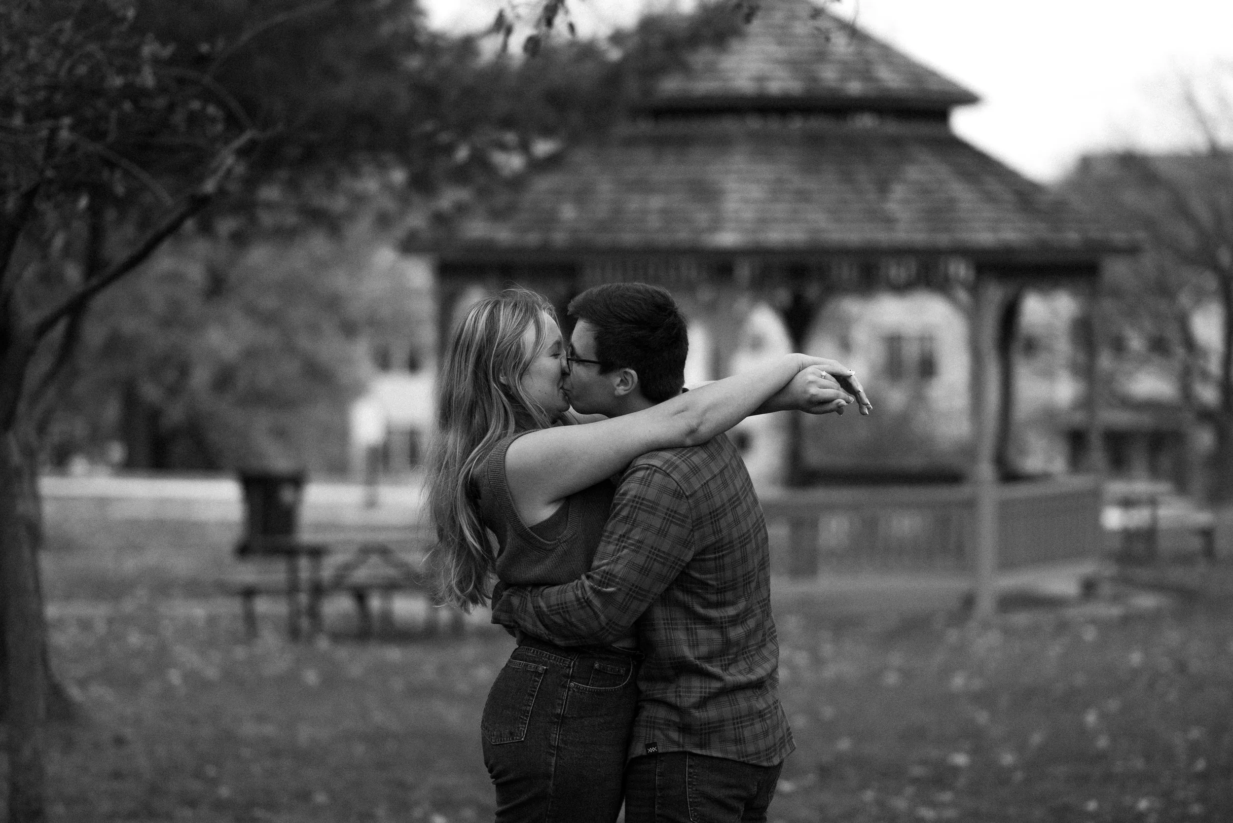 A couple kissing in a park, with the woman wrapping her arms around the man's neck and the man holding her waist, in front of a gazebo.