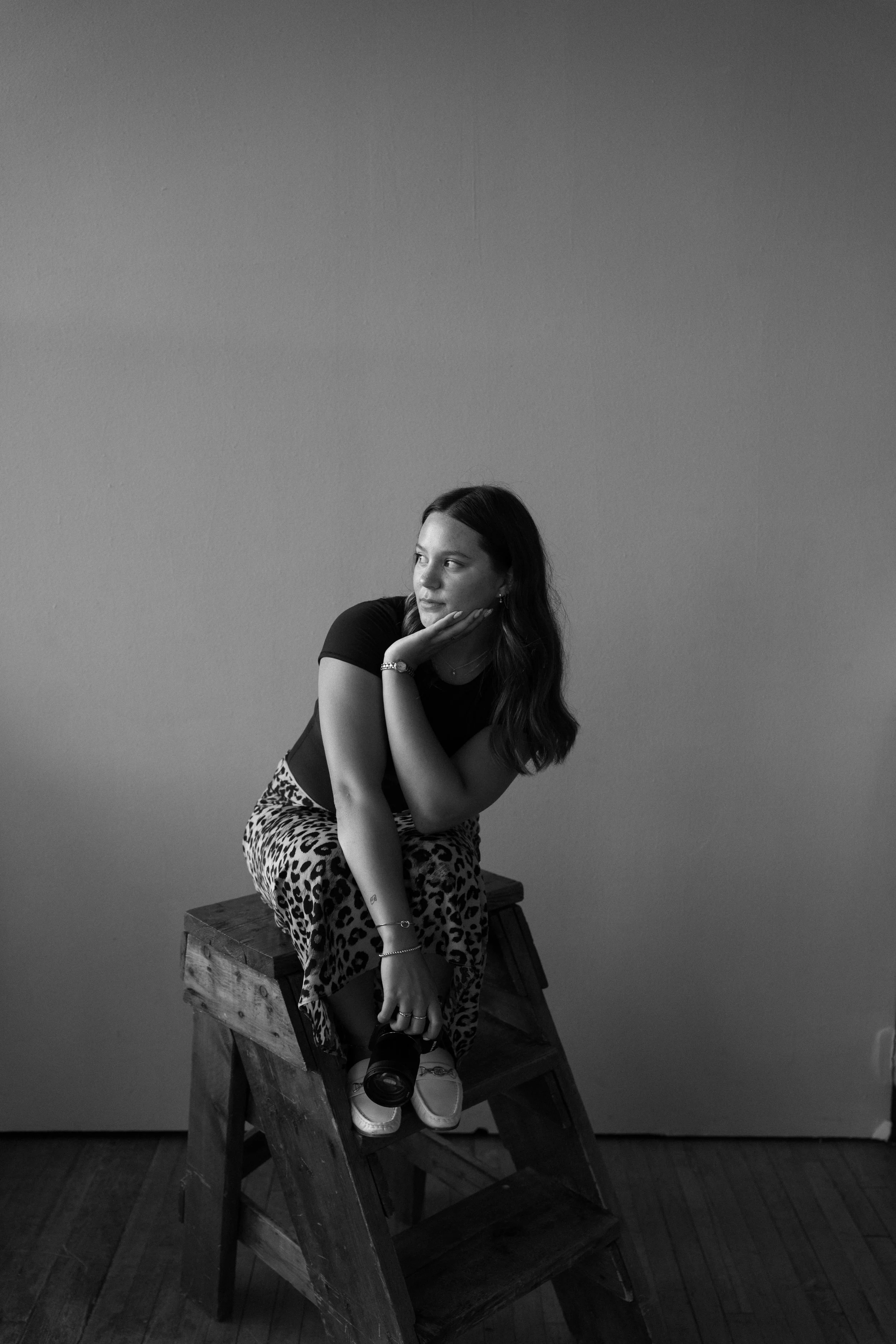 A woman with long dark hair sitting on a wooden ladder against a plain wall, holding a camera and resting her chin on her hand, in black and white.