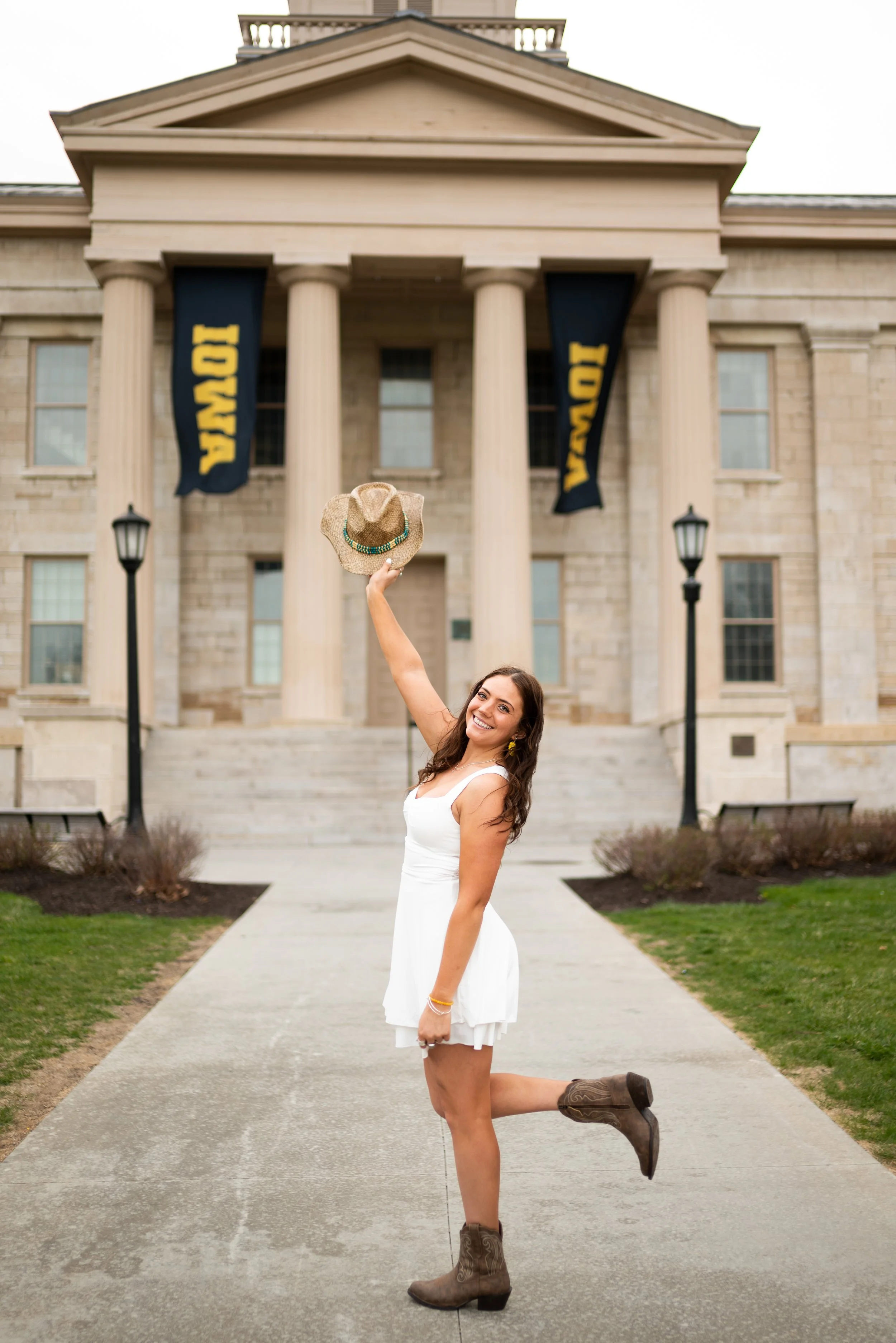 A young woman in a white dress and cowboy boots happily posing outside a college building with flags that read 'IOWA'. She is holding a cowboy hat in the air and standing on a sidewalk.