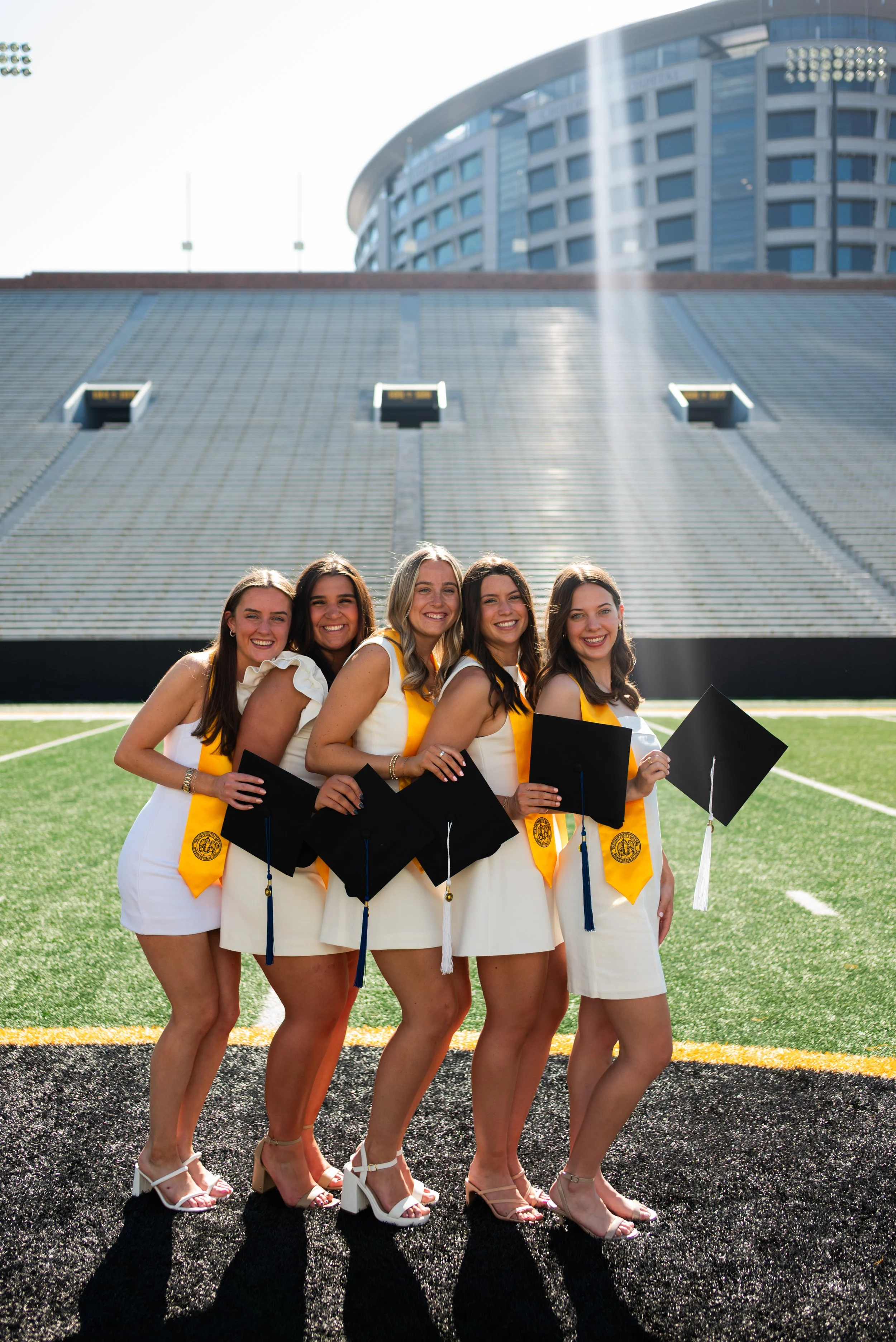 Group of five young women in white dresses with yellow stoles holding diplomas and posing for a graduation photo on a football field in an outdoor stadium.