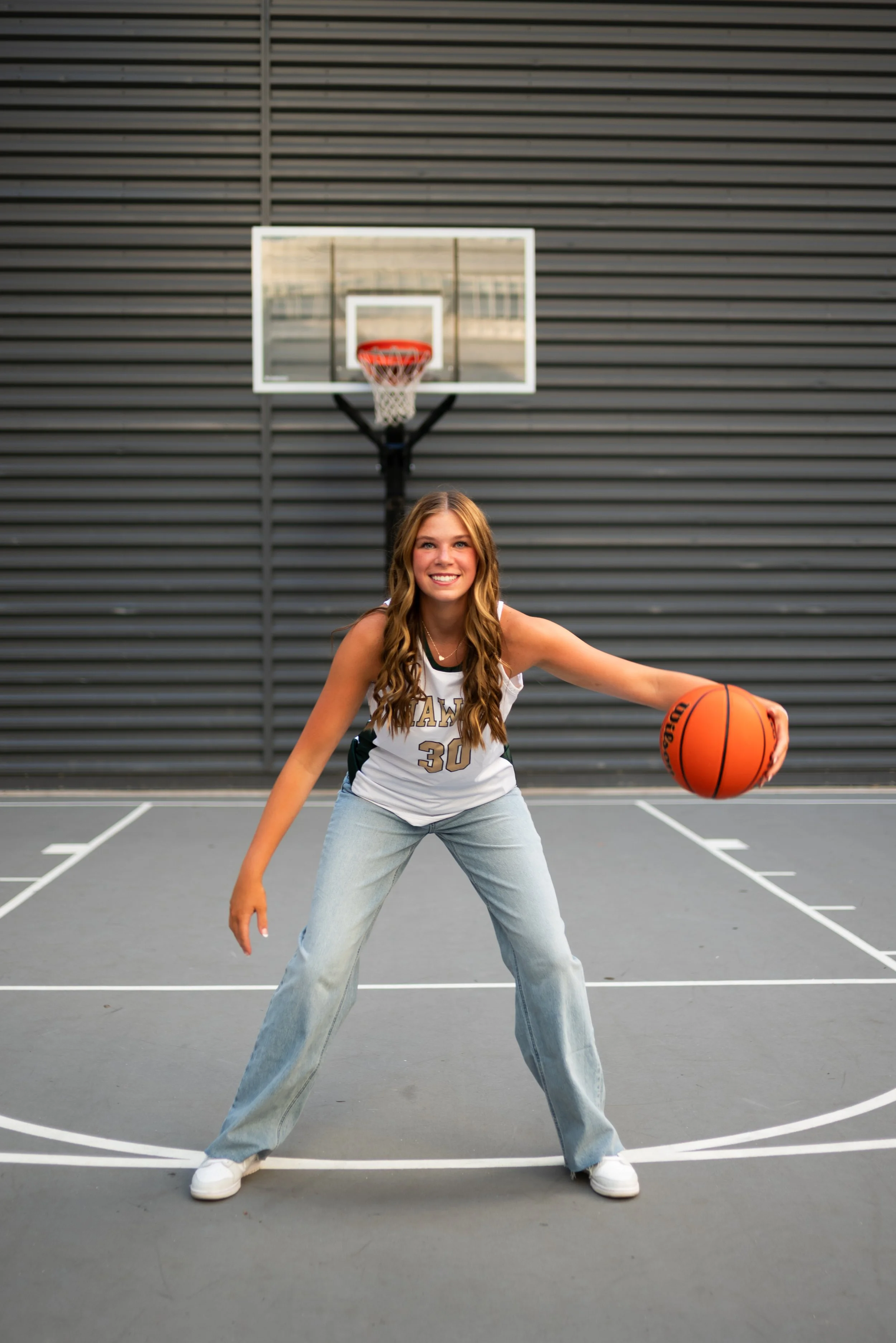 Young woman on a basketball court holding a basketball, smiling, with a basketball hoop behind her.