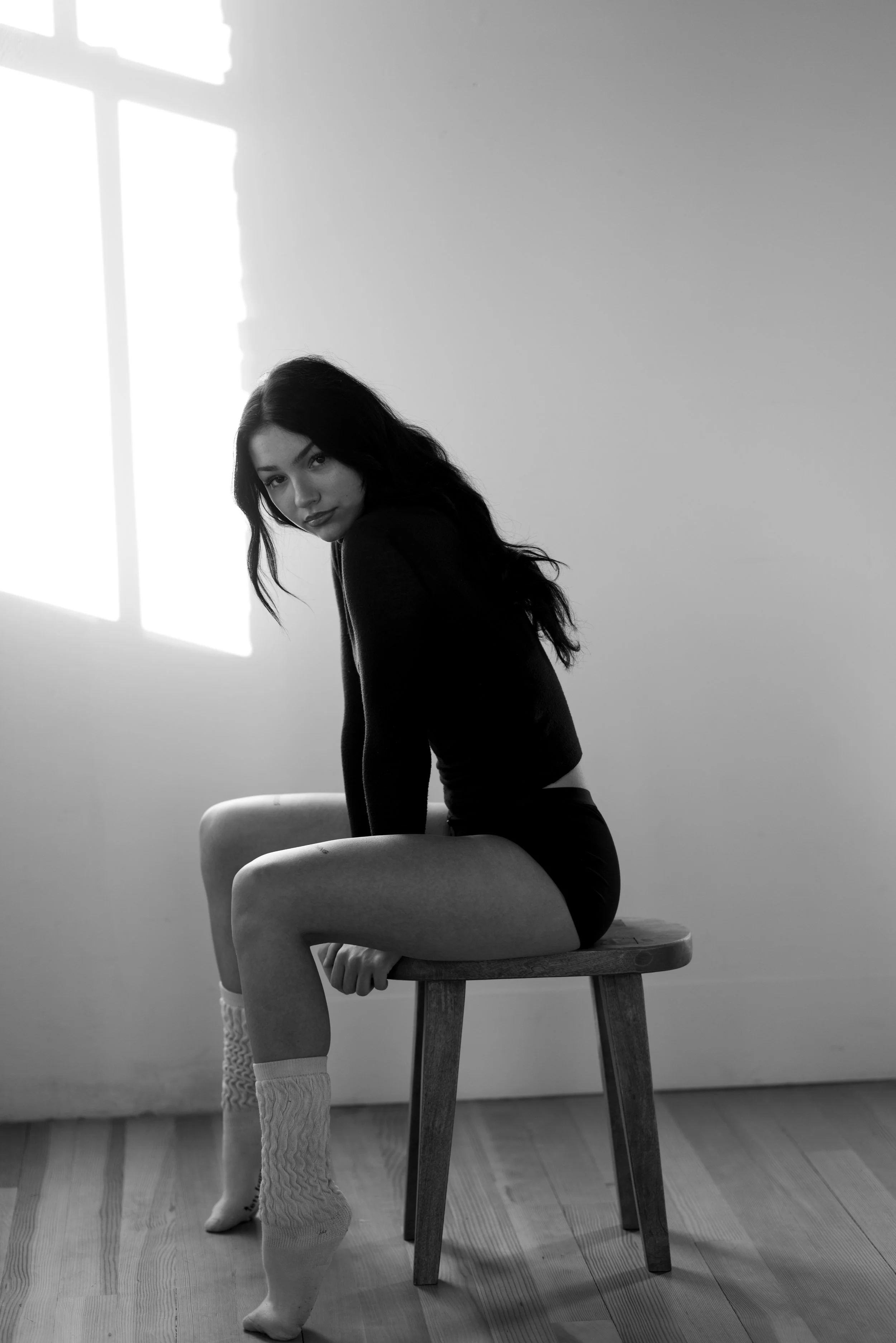 A young woman with long dark hair sitting on a wooden stool in a room with sunlight coming through a window, wearing a dark top, shorts, and cable-knit socks.