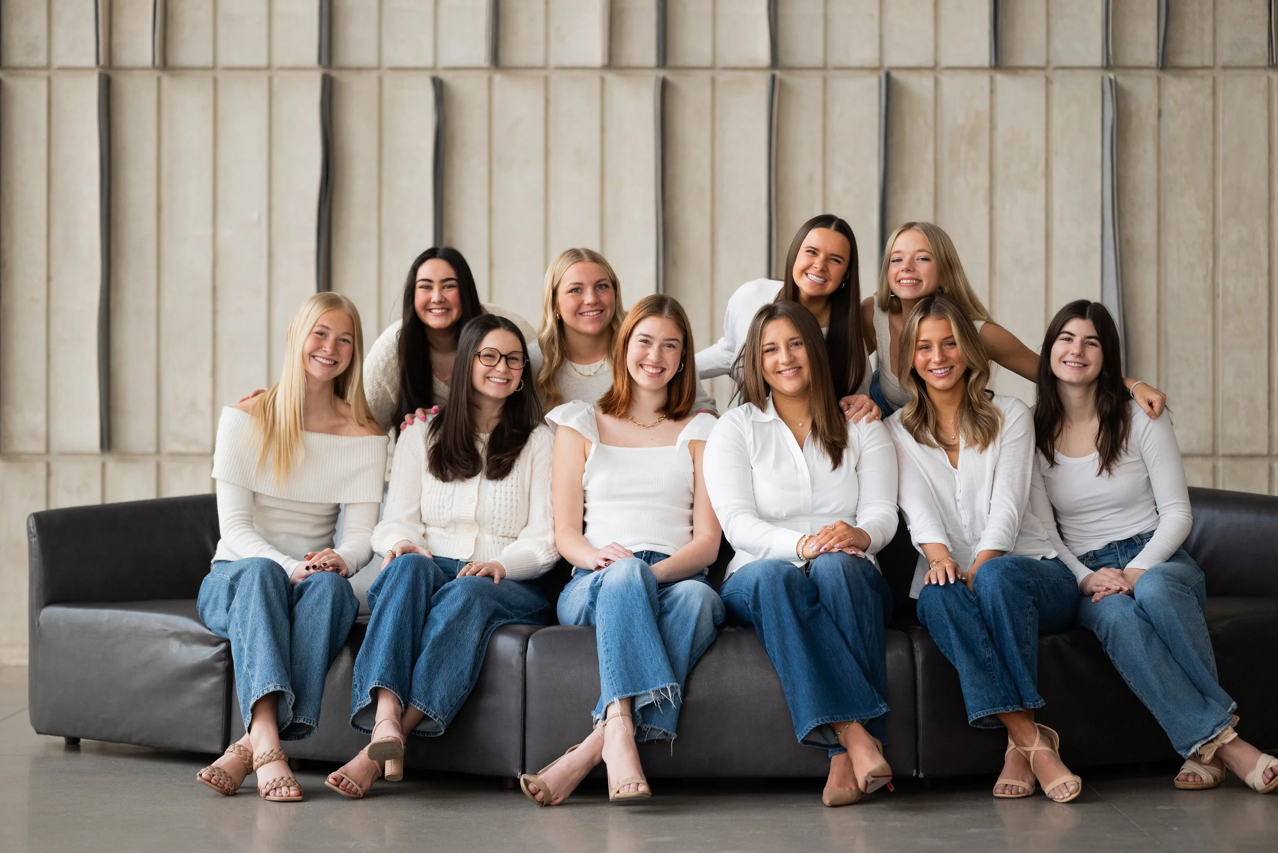 A group of ten young women posing together indoors, sitting and standing on a black couch, dressed in white tops and jeans, smiling for the camera.