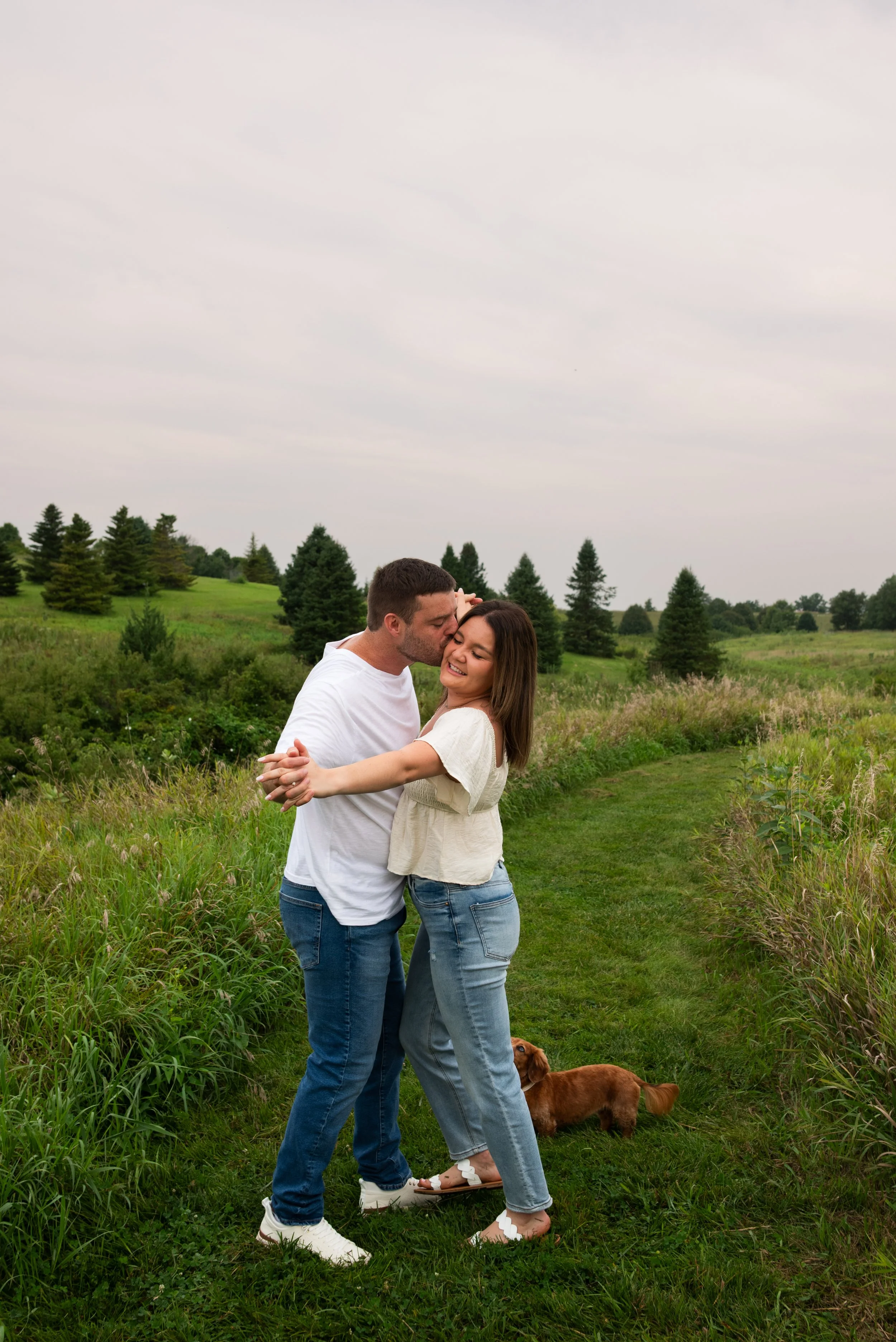 A couple hugging and smiling in a grassy field with a small brown dog nearby, surrounded by greenery and trees under a cloudy sky.