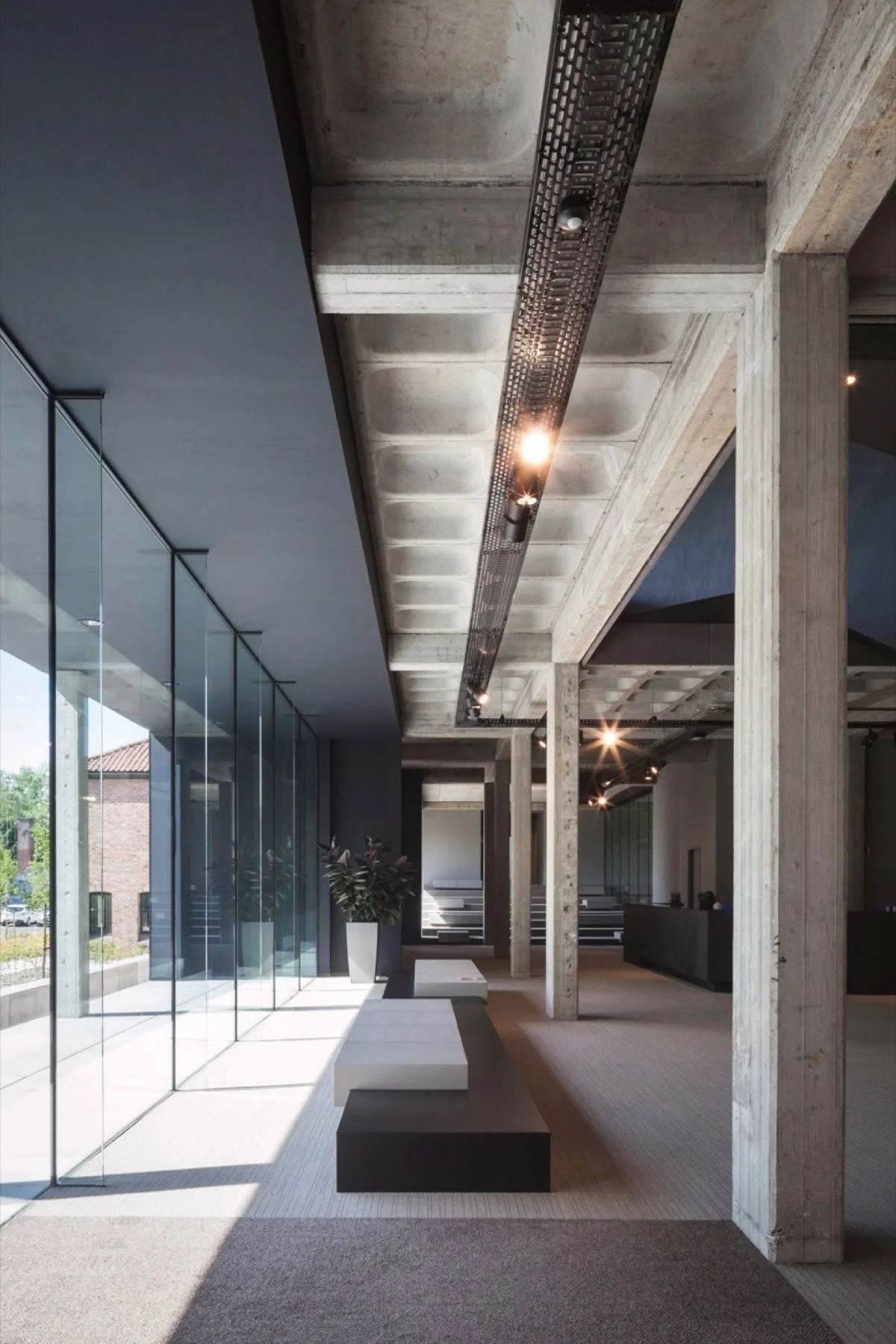 Interieur van een modern gebouw met grote glasspanelen, stoelen, planten, betonnen kolommen en een benzine-achtige markering aan het plafond.