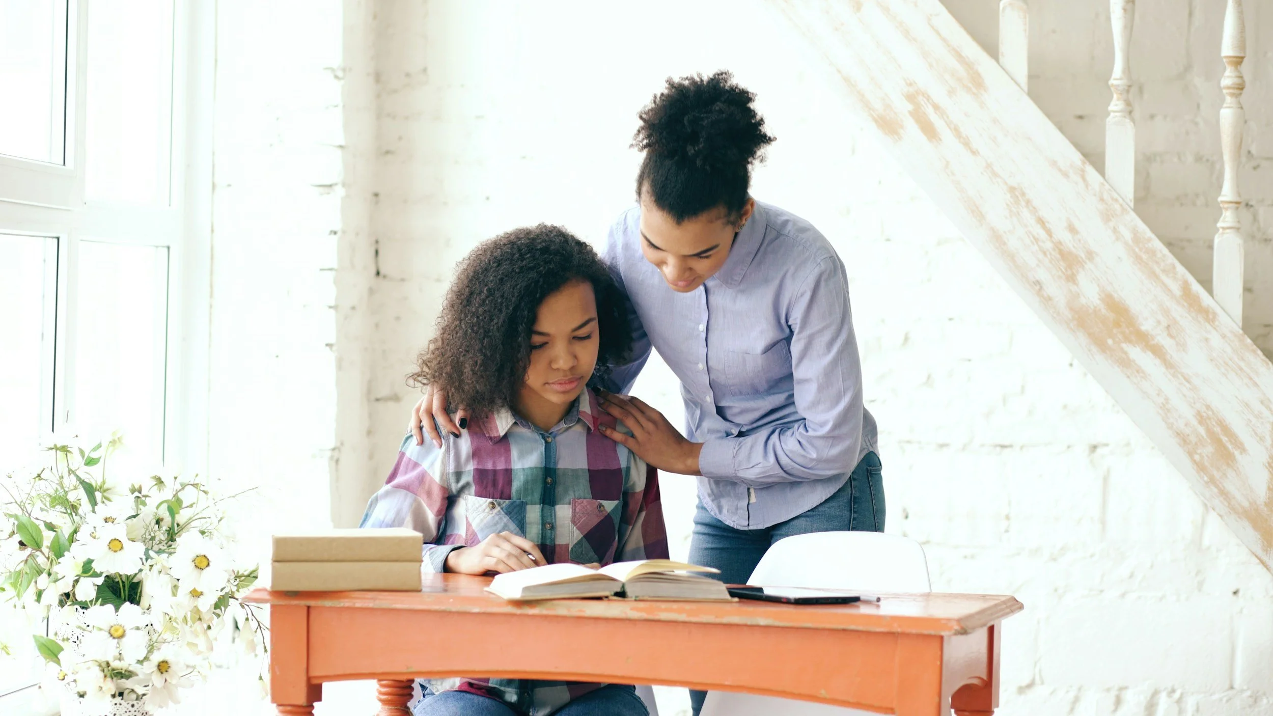 Two women, one sitting at a table and the other standing beside her, comforting her while they look at a book in a bright room with white brick walls.