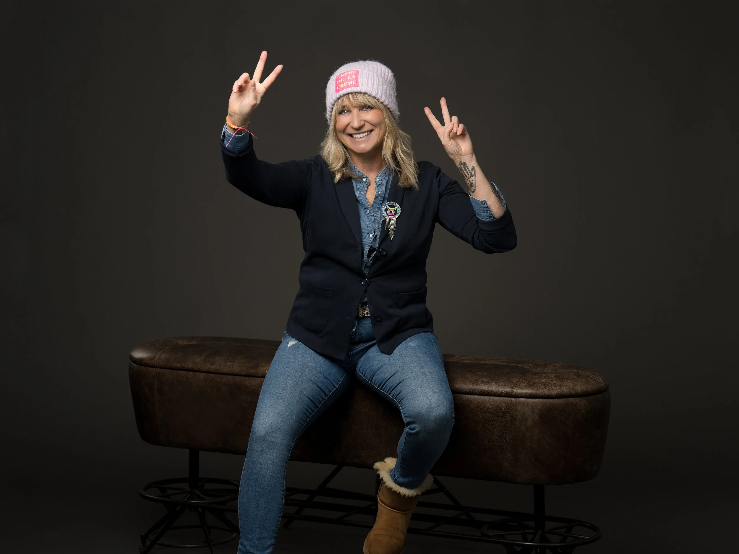 Eine lächelnde Frau in Jeans, schwarzer Jacke, grauem Beanie und braunen Stiefeln sitzt auf einer braunen Couch vor dunklem Hintergrund und macht Peace-Zeichen mit beiden Händen.