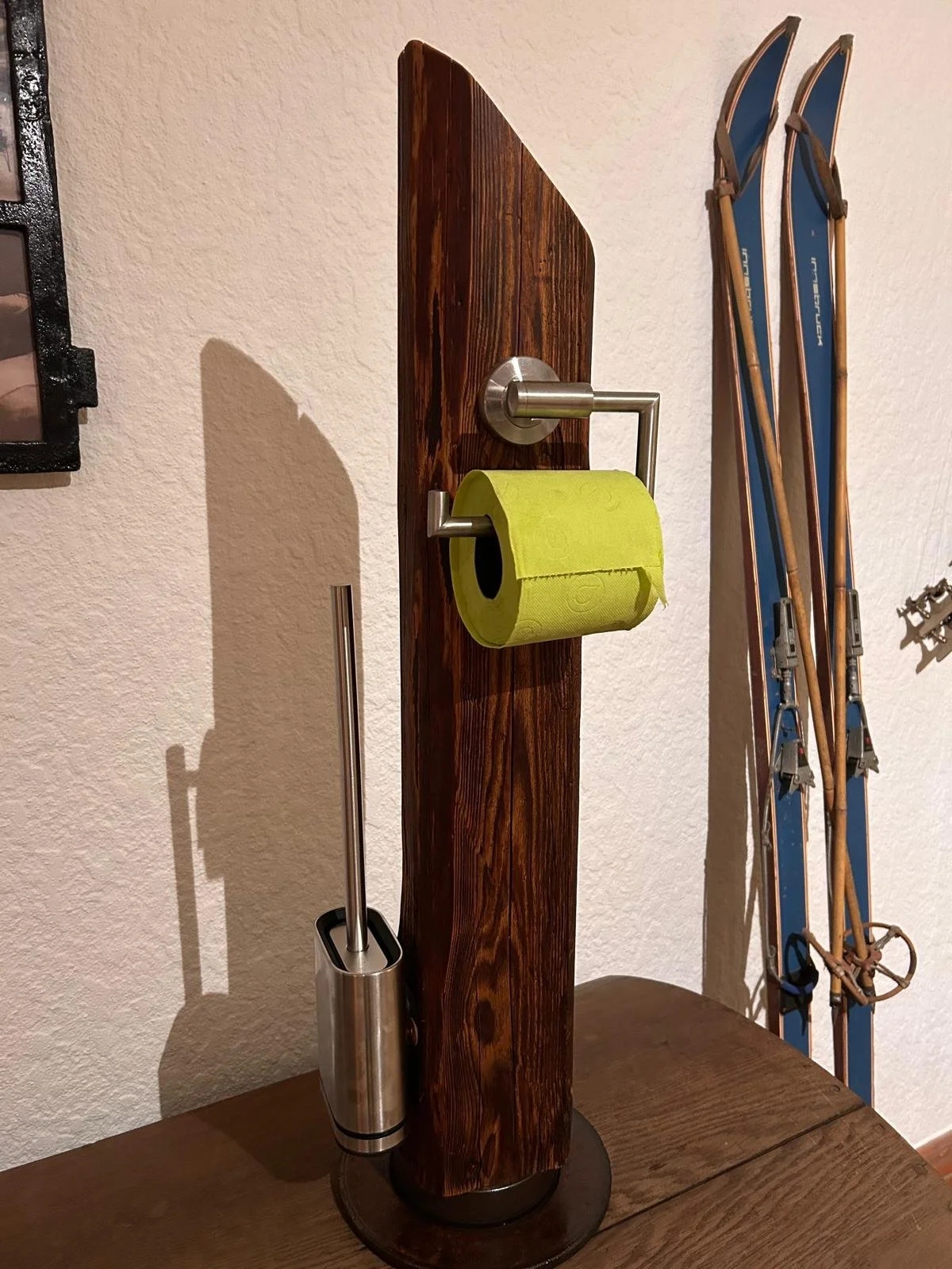 Ein moderner Toilettenpapierhalter aus Edelstahl an einem Holzständer mit einer Rolle grünem Toilettenpapier, daneben ein WC-Bürstenset, auf der rechten Seite Skistöcke.