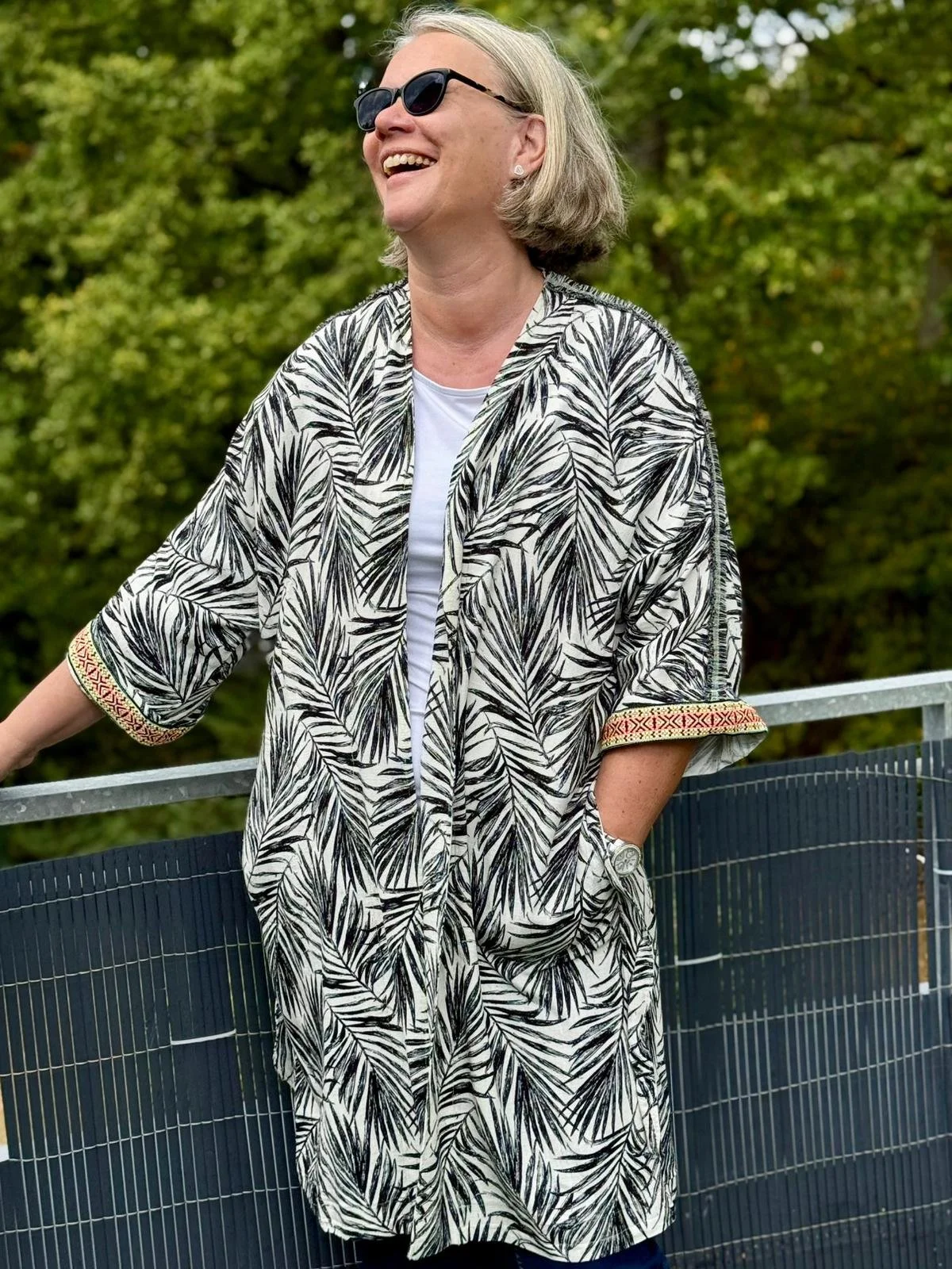 Eine lachende ältere Frau mit Sonnenbrille in einem zigarren Muster-Kleid im Freien vor grünen Bäumen.