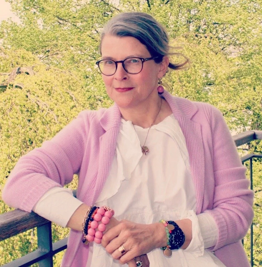 Frau mit Brille in einer rosa Jacke sitzt auf einem Balkon vor grünen Bäumen.