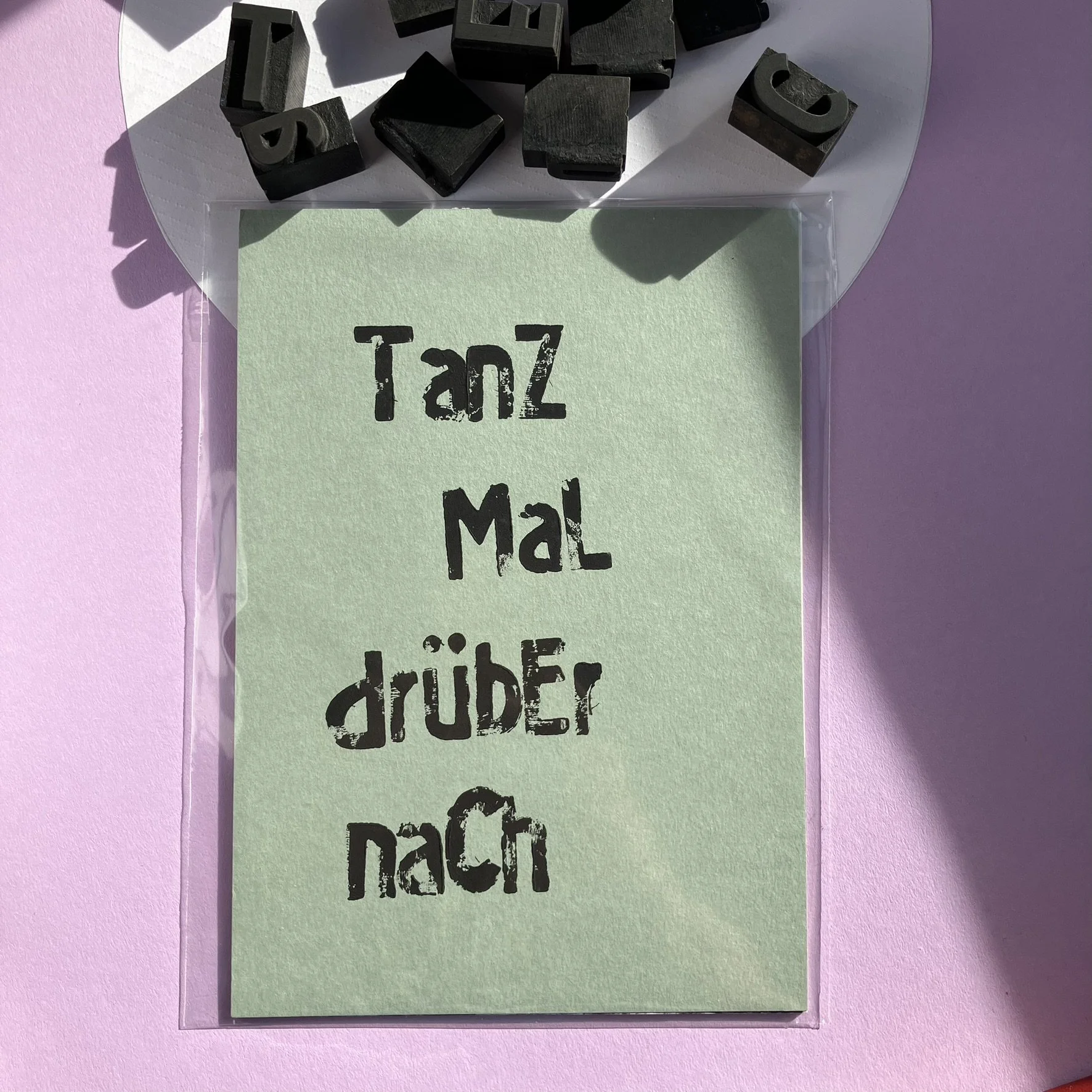 Ein grünes Post-it mit der Aufschrift "Tanz Mal drüben nach" in schwarzen Buchstaben, umgeben von schwarzen Buchstabenstempeln auf einer weißen Unterlage, alles auf einem rosa Hintergrund.