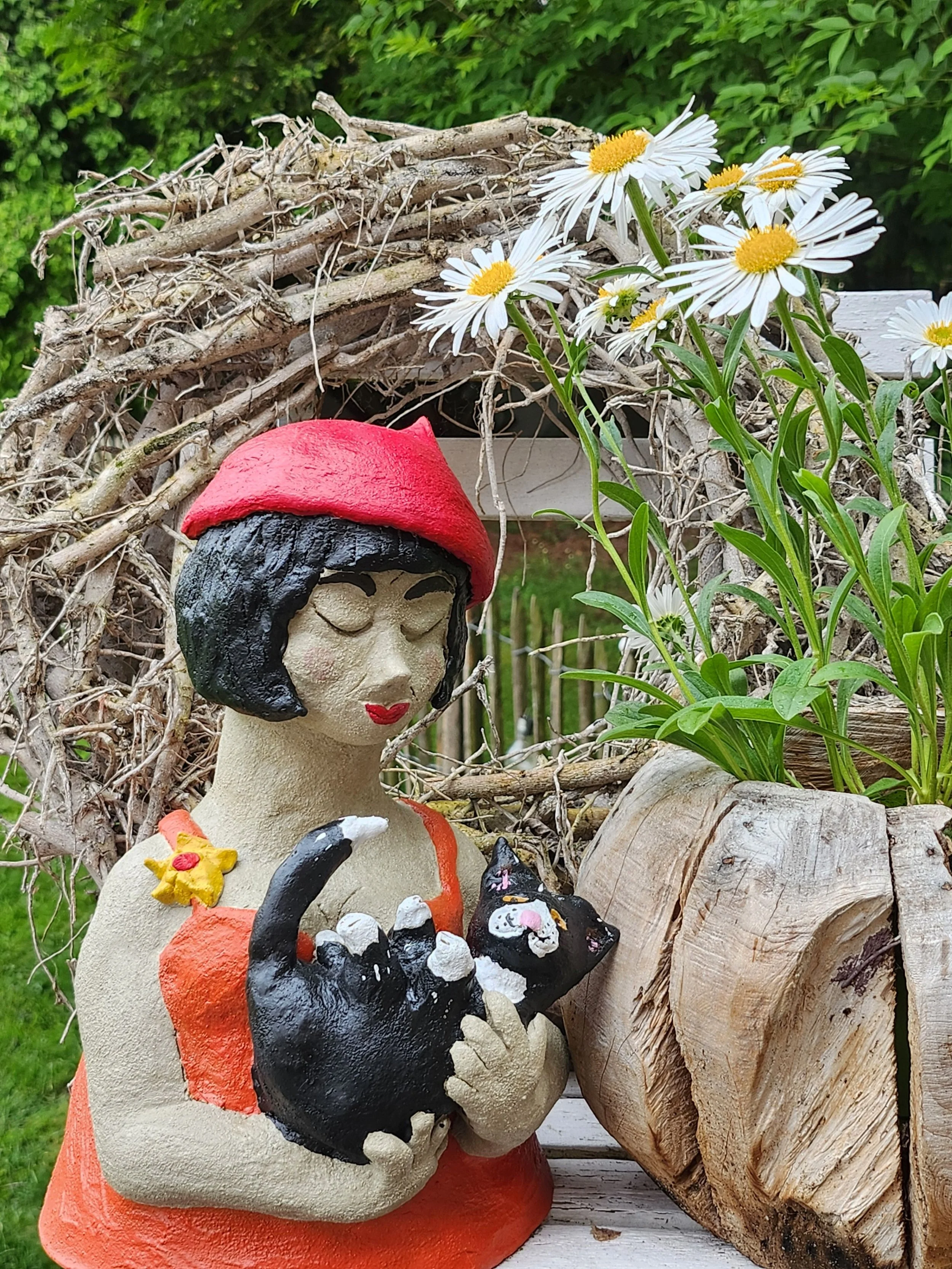 Eine Skulptur einer Frau mit schwarzen Haaren, rotem Hut und rotem Kleid, die eine schwarze Katze hält, davor Blumen und ein Zweig
