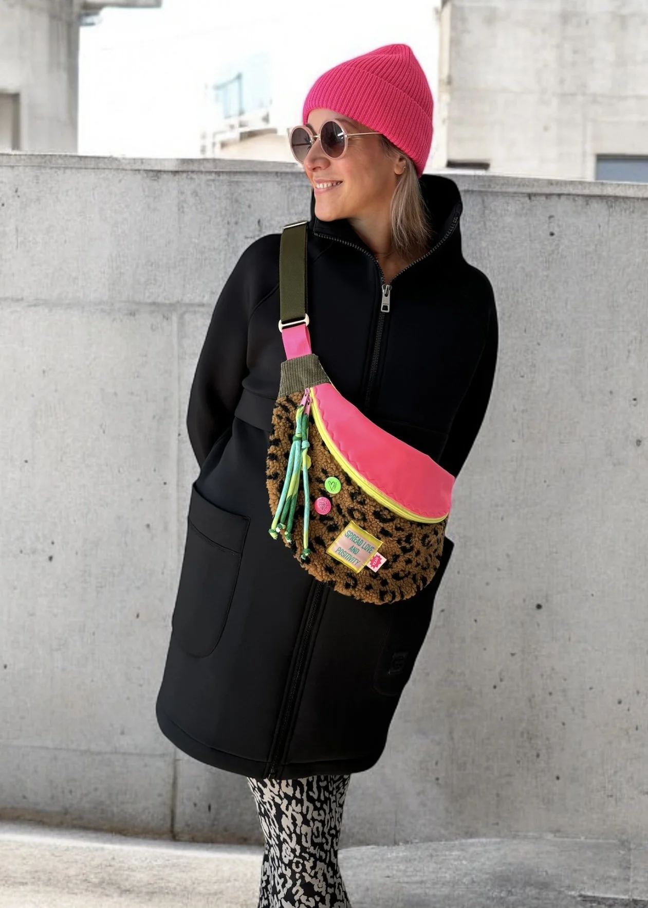 Eine lachende Frau mit Sonnenbrille, pinkem Mütze, schwarzer Jacke und Leoparden-Musterhose, trägt eine Umhängetasche mit bunten Pins und Anhängern, steht vor einer Betonmauer.