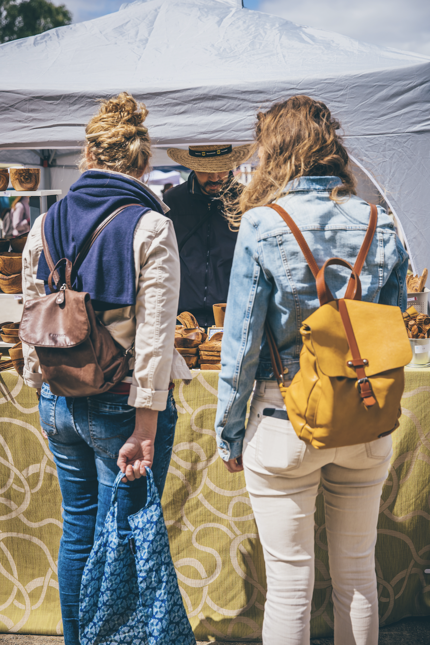 Zwei Frauen sprechen mit einem Mann an einem Marktstand mit Holzprodukten im Freien.