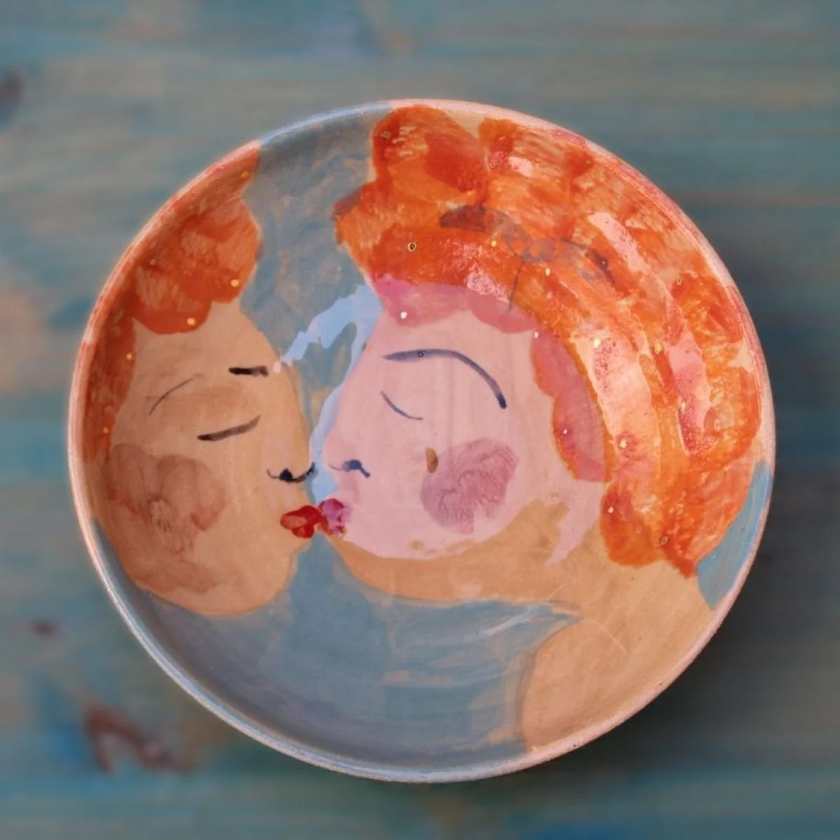 Kunstvoll bemalte Schüssel mit zwei Frauen, die sich küssen, auf orangefarbenem Hintergrund, auf einem blauen Holztisch.