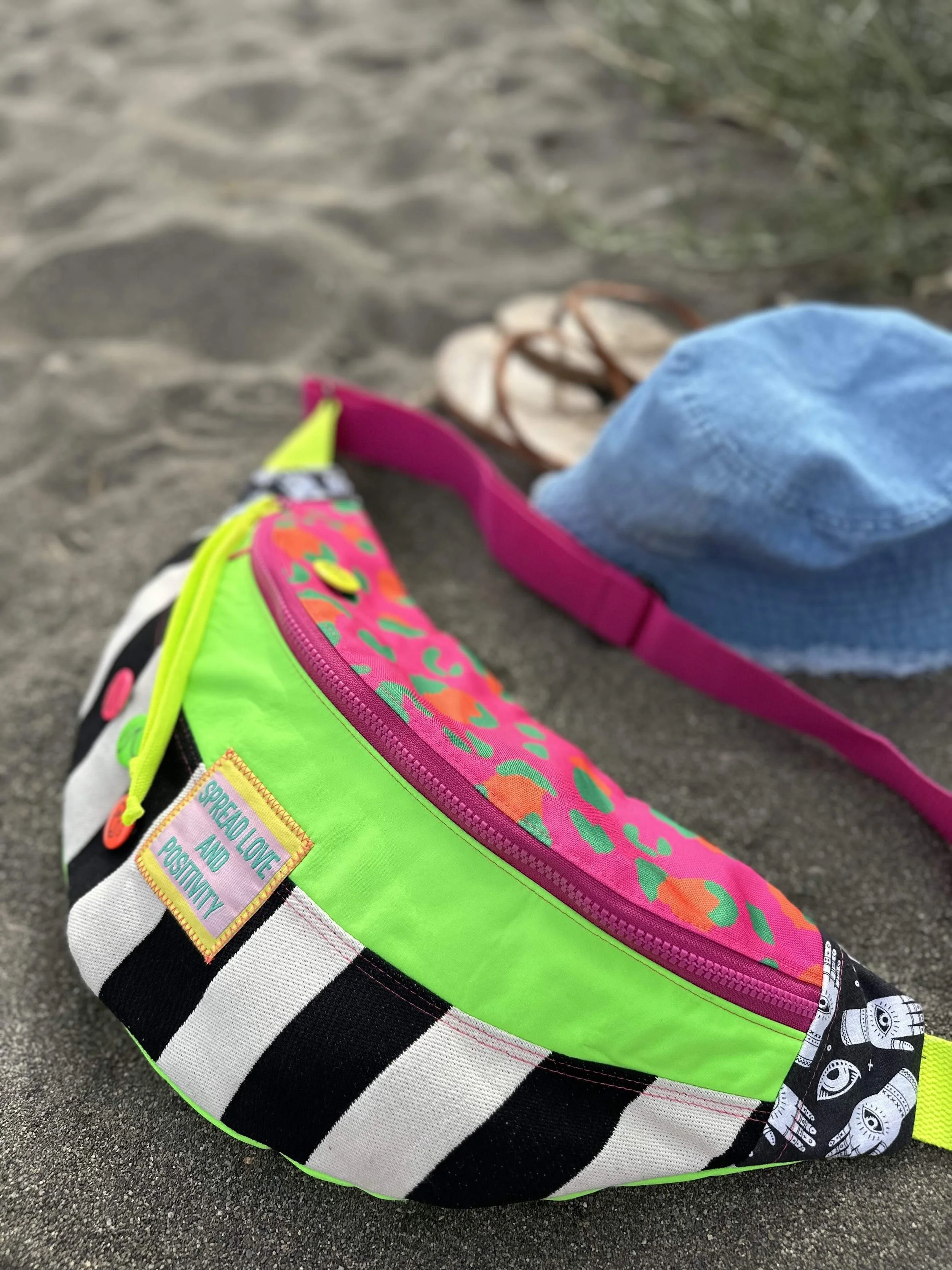 Bunte Gürteltasche mit Streifenmuster, leuchtenden Farben und Aufkleber, die eine positive Botschaft tragen, auf Sand vor einer Strandkulisse, neben einer blauen Mütze und Sandalen