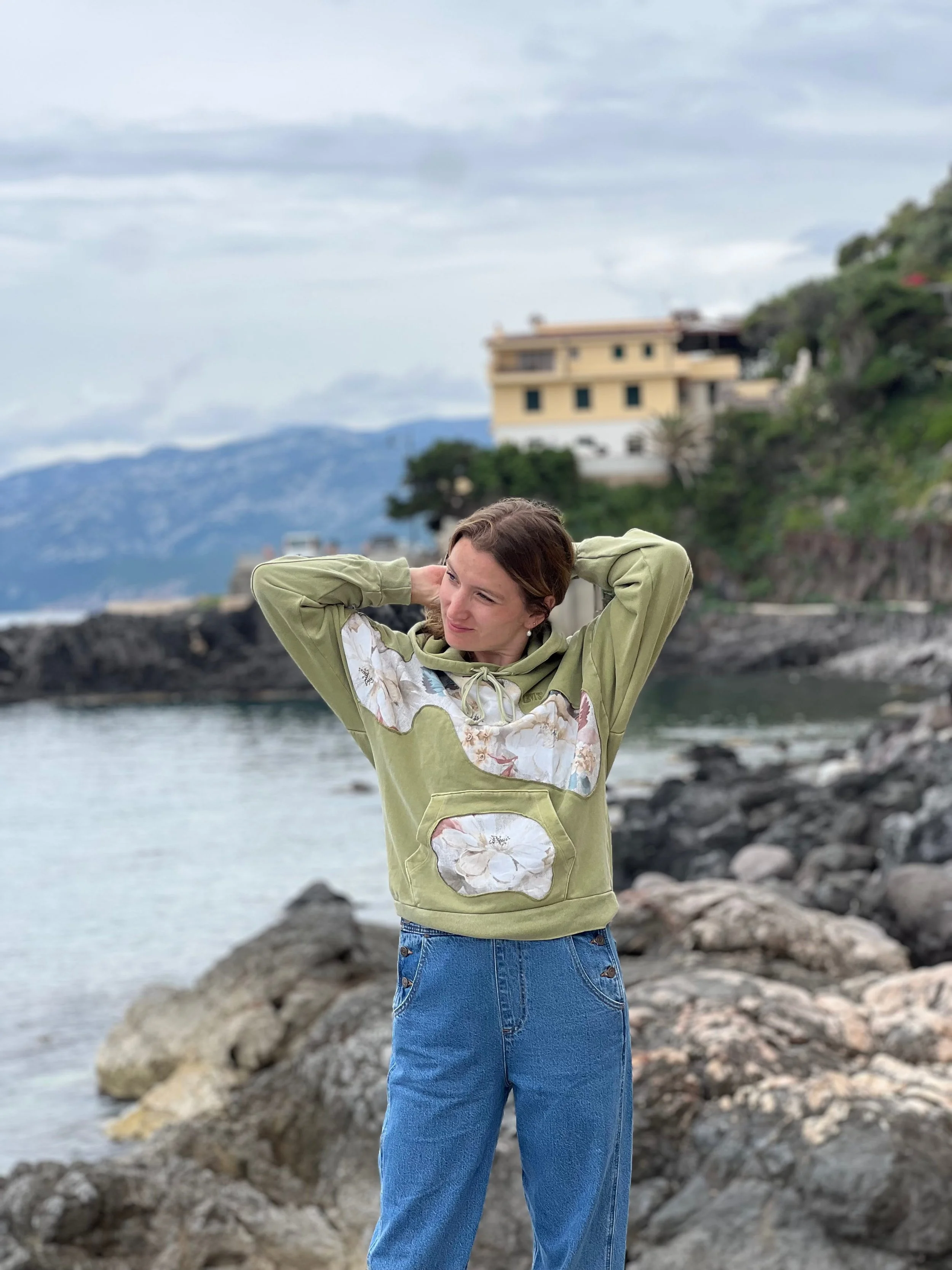 Junge Frau mit braunen Haaren in grünem Hoodie mit weißen Blumenmustern und blauen Jeans am Felsen an einem See mit Häusern und Bergen im Hintergrund.