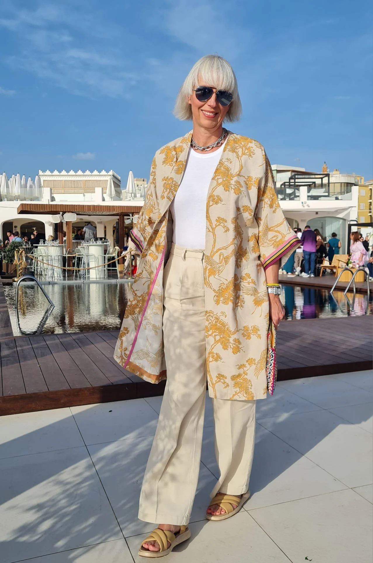 Eine ältere Frau mit weißen Haaren in sommerlicher Kleidung und Sonnenbrille steht an einem Pool im Freien, umgeben von Menschen bei schönem Wetter.
